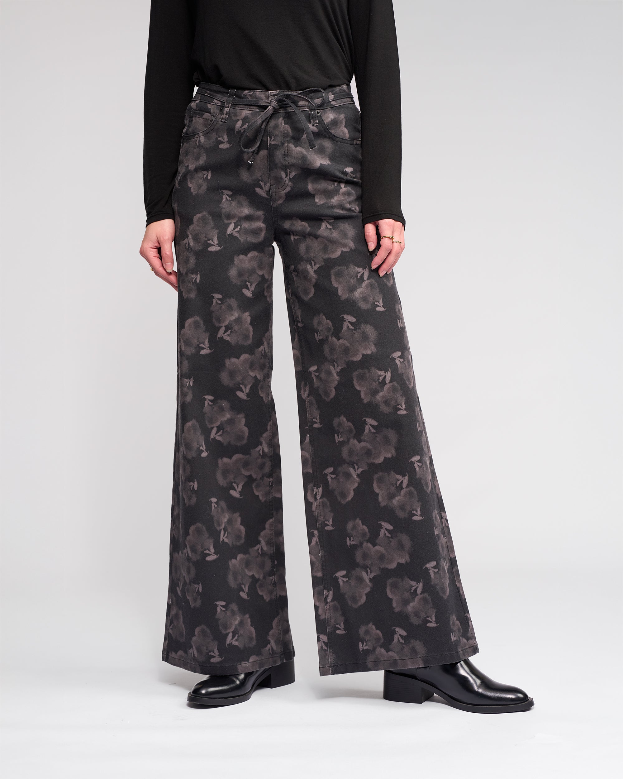 Against a plain light background, a person models the Knewe Label Goldie Jean - Echo: wide-leg, dark floral denim pants. Their black long-sleeve top and black heeled shoes are visible, but their head is cropped out of the image.