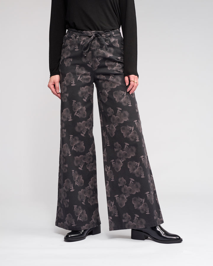 Against a plain light background, a person models the Knewe Label Goldie Jean - Echo: wide-leg, dark floral denim pants. Their black long-sleeve top and black heeled shoes are visible, but their head is cropped out of the image.