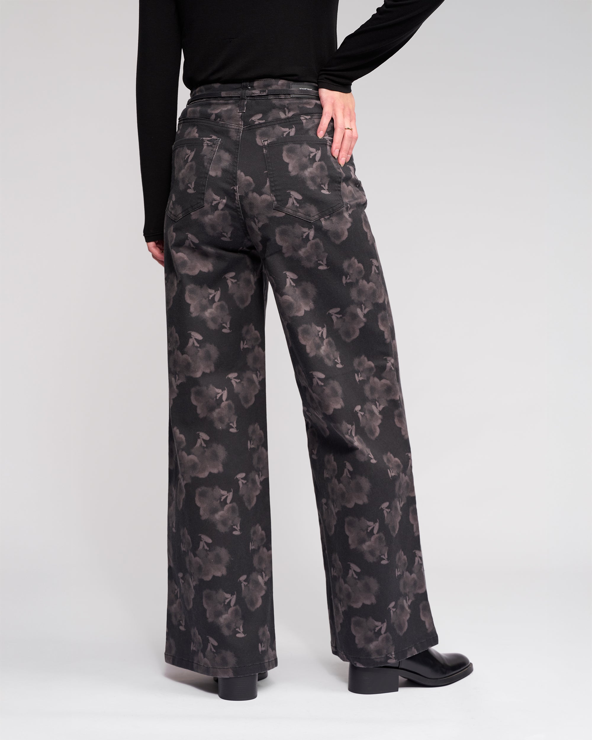 A person wearing Knewe Label's Goldie Jean - Echo, high-waisted wide-leg jeans, with a black long-sleeve top. The photo captures the back of the floral denim as the person's hand rests on their hip.