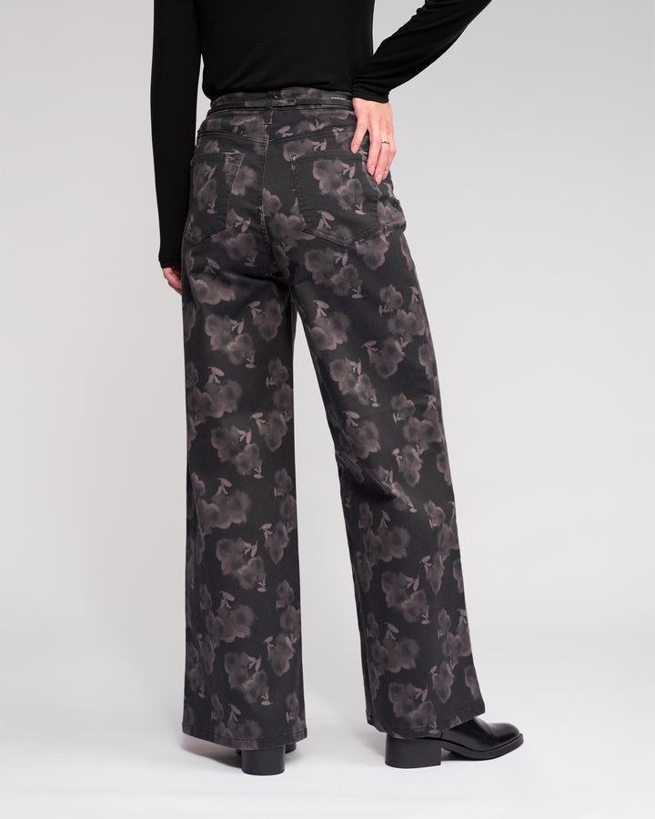 A person wearing Knewe Label's Goldie Jean - Echo, high-waisted wide-leg jeans, with a black long-sleeve top. The photo captures the back of the floral denim as the person's hand rests on their hip.