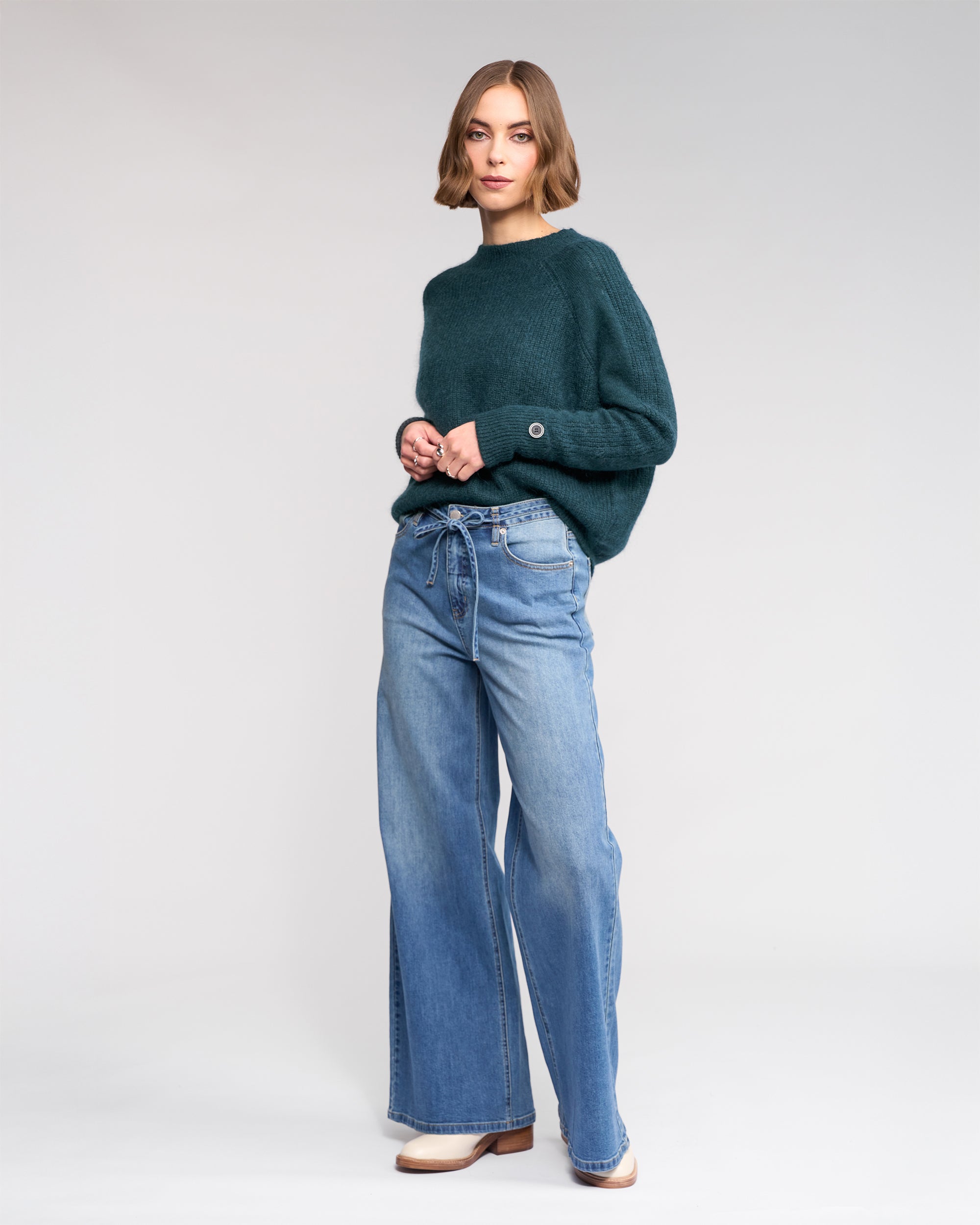A woman with a bob haircut wears a dark green sweater and Knewe Label’s Goldie Jean in washed denim, featuring a wide-leg fit and tie belt, while standing against a plain light background.