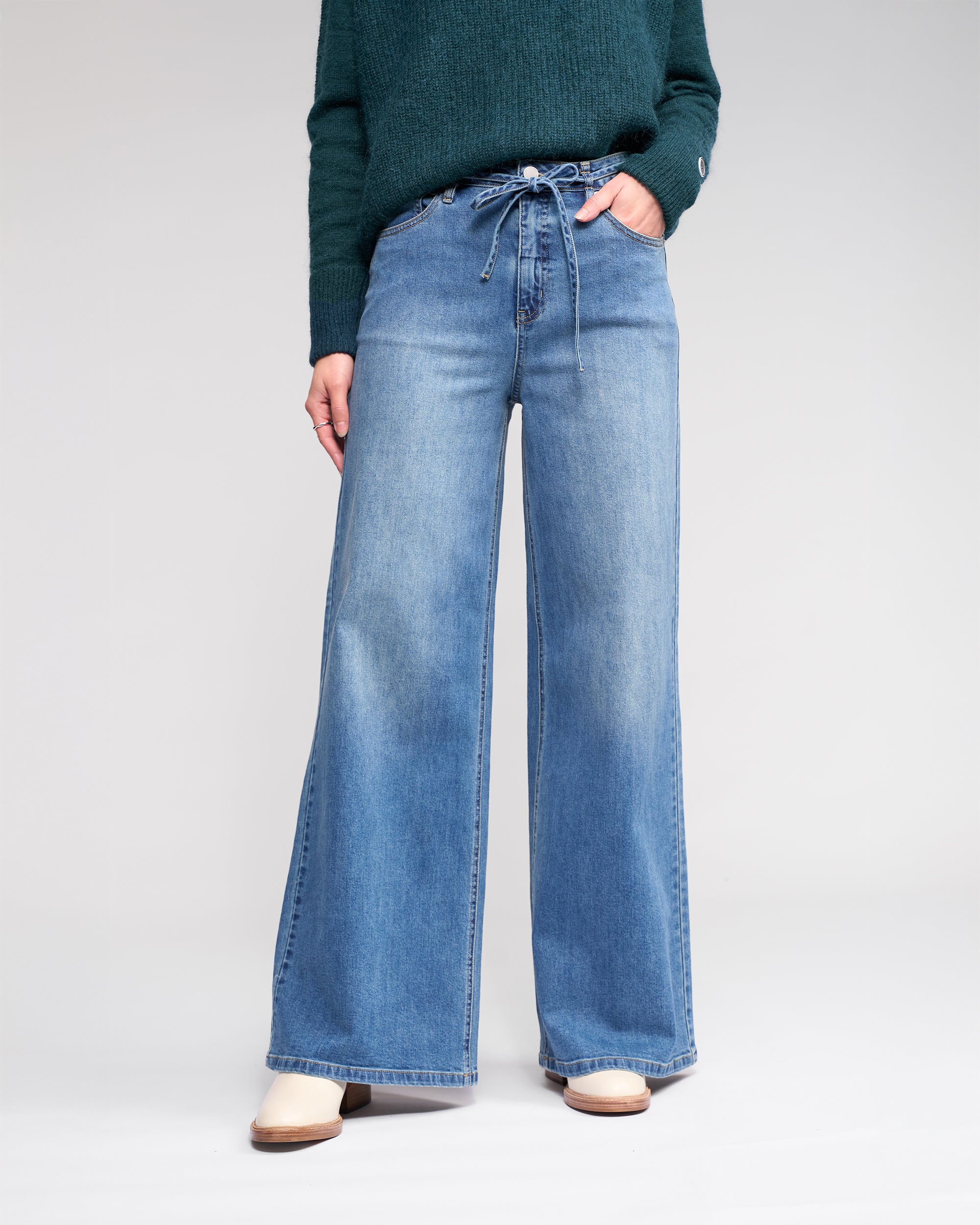 Wearing Knewe Label's Goldie Jean in washed denim with a tie belt, paired with a dark green sweater and cream shoes, the person stands against a plain light background, face not visible.