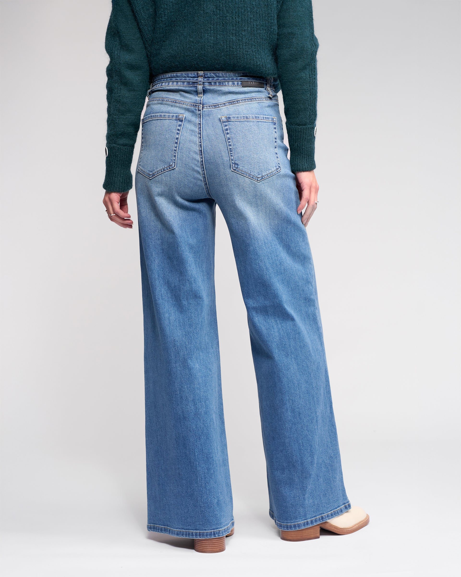 Someone stands facing away, wearing Knewe Label’s Goldie Jean in washed denim—high-waisted, wide-leg jeans with a denim tie belt—paired with a dark green sweater and light shoes, all set against a plain background.
