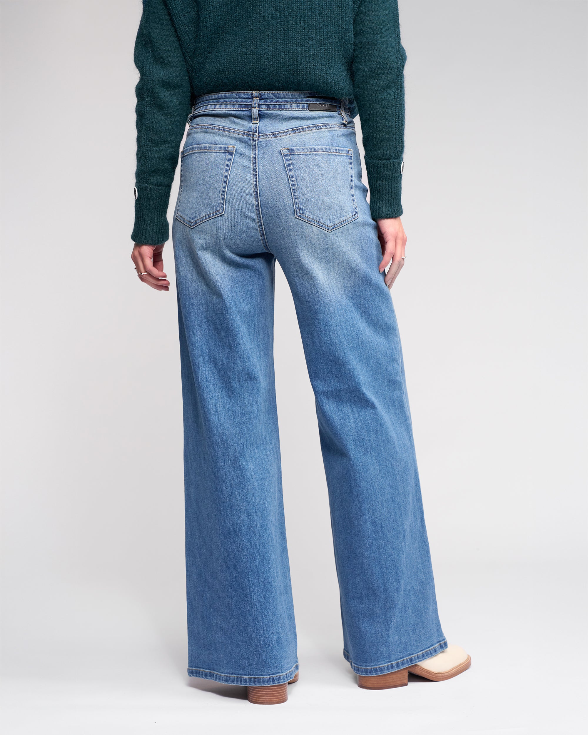 Someone stands facing away, wearing Knewe Label’s Goldie Jean in washed denim—high-waisted, wide-leg jeans with a denim tie belt—paired with a dark green sweater and light shoes, all set against a plain background.
