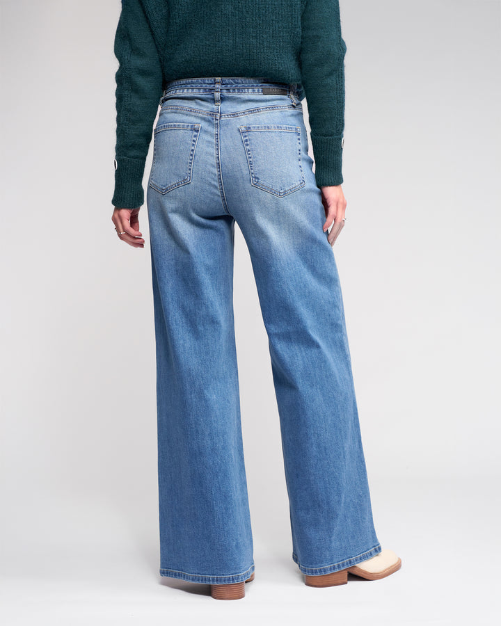 Someone stands facing away, wearing Knewe Label’s Goldie Jean in washed denim—high-waisted, wide-leg jeans with a denim tie belt—paired with a dark green sweater and light shoes, all set against a plain background.