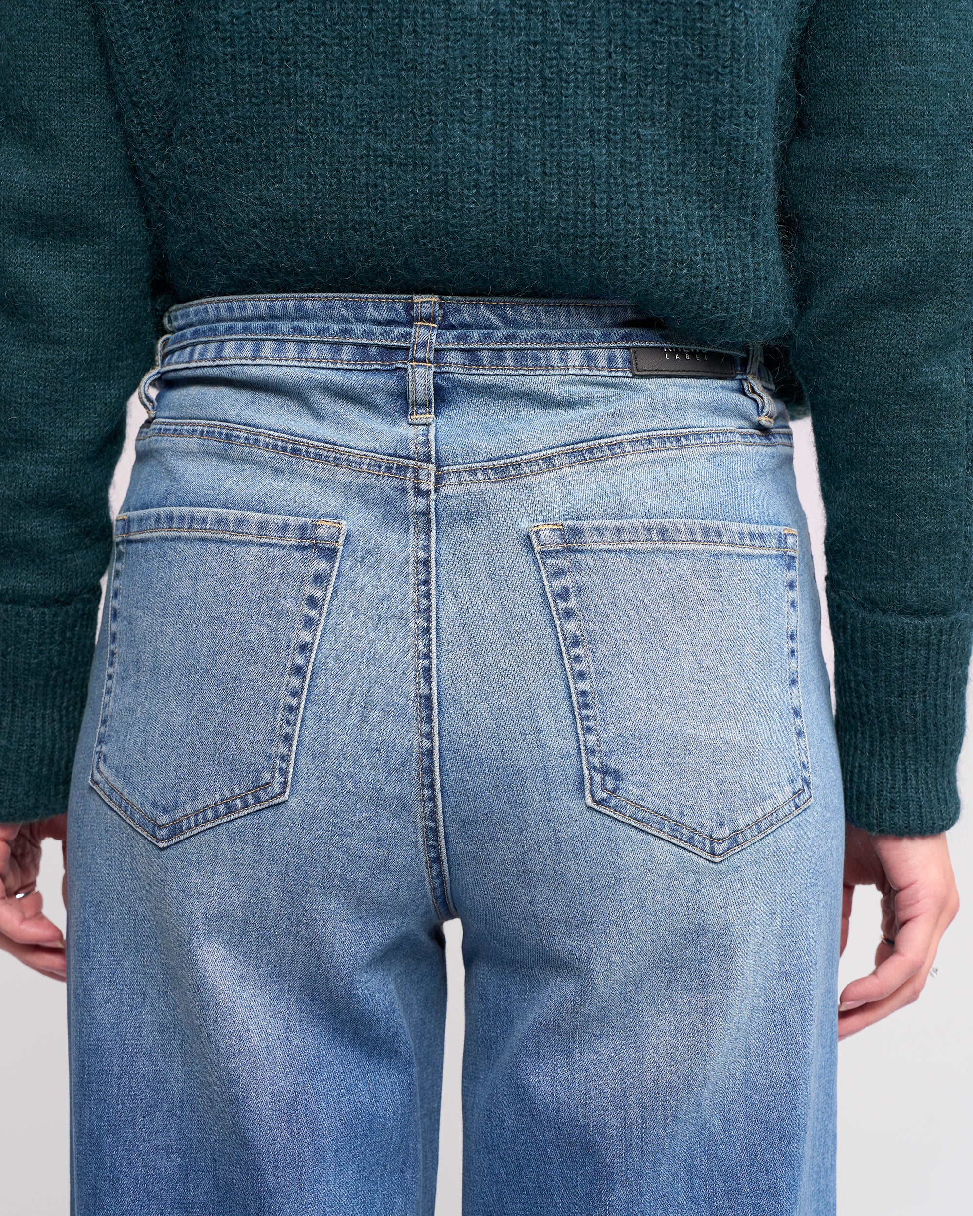 A person is shown from the back wearing Knewe Label's Goldie Jean in washed denim—light blue, high-waisted, wide-leg with visible back pockets and belt loops—paired with a dark green knitted sweater, hands relaxed at their sides.