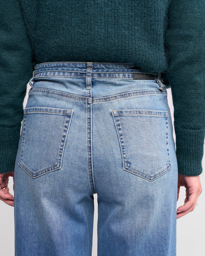 A person is shown from the back wearing Knewe Label's Goldie Jean in washed denim—light blue, high-waisted, wide-leg with visible back pockets and belt loops—paired with a dark green knitted sweater, hands relaxed at their sides.