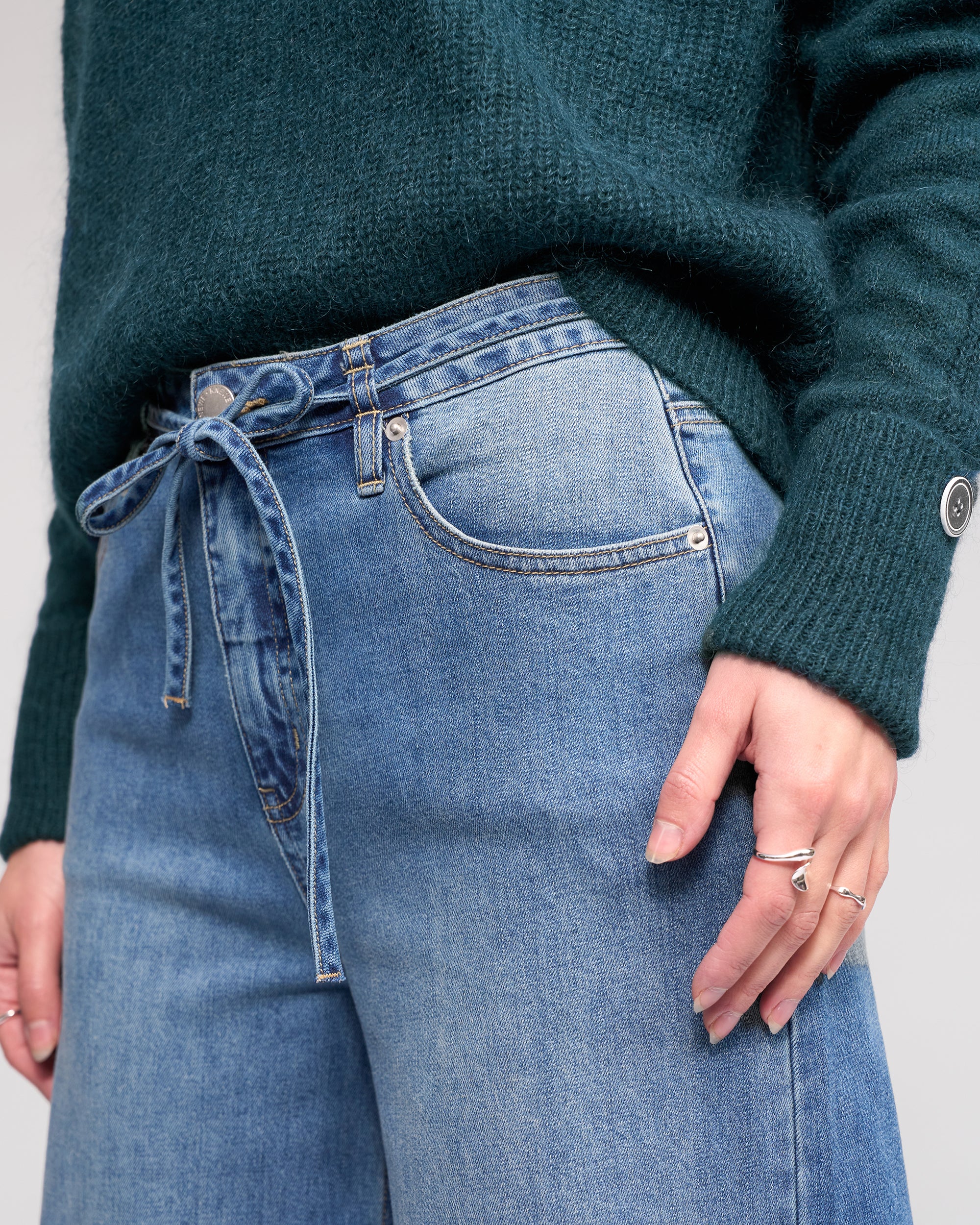 Wearing the Knewe Label’s Goldie Jean in washed denim, styled with a denim tie belt in a bow and paired with a green sweater. Only the torso and upper legs are shown as a ring-adorned hand rests near the pocket.