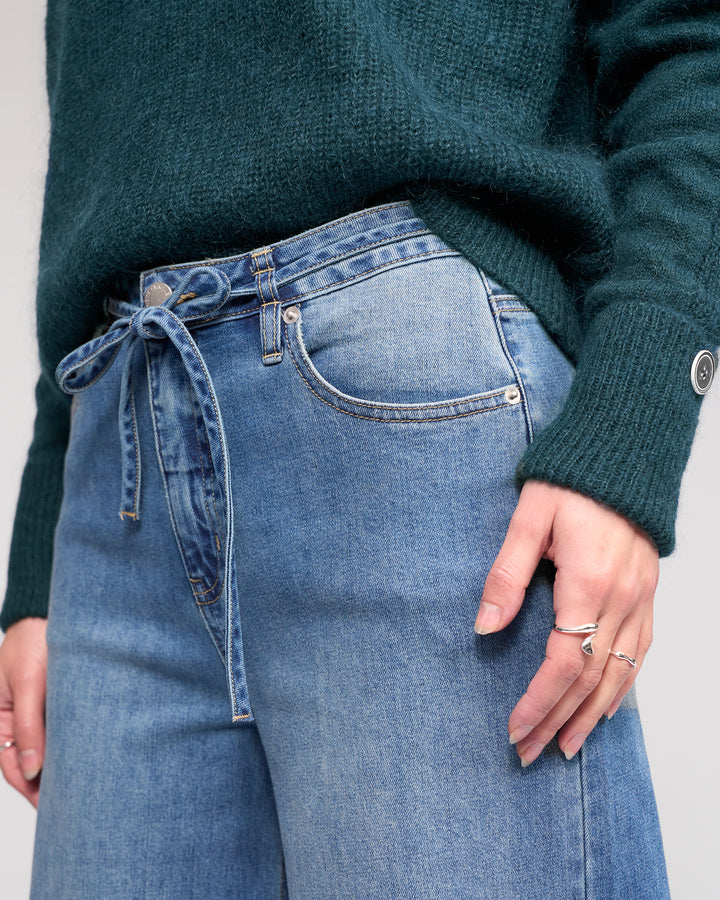 Wearing the Knewe Label’s Goldie Jean in washed denim, styled with a denim tie belt in a bow and paired with a green sweater. Only the torso and upper legs are shown as a ring-adorned hand rests near the pocket.