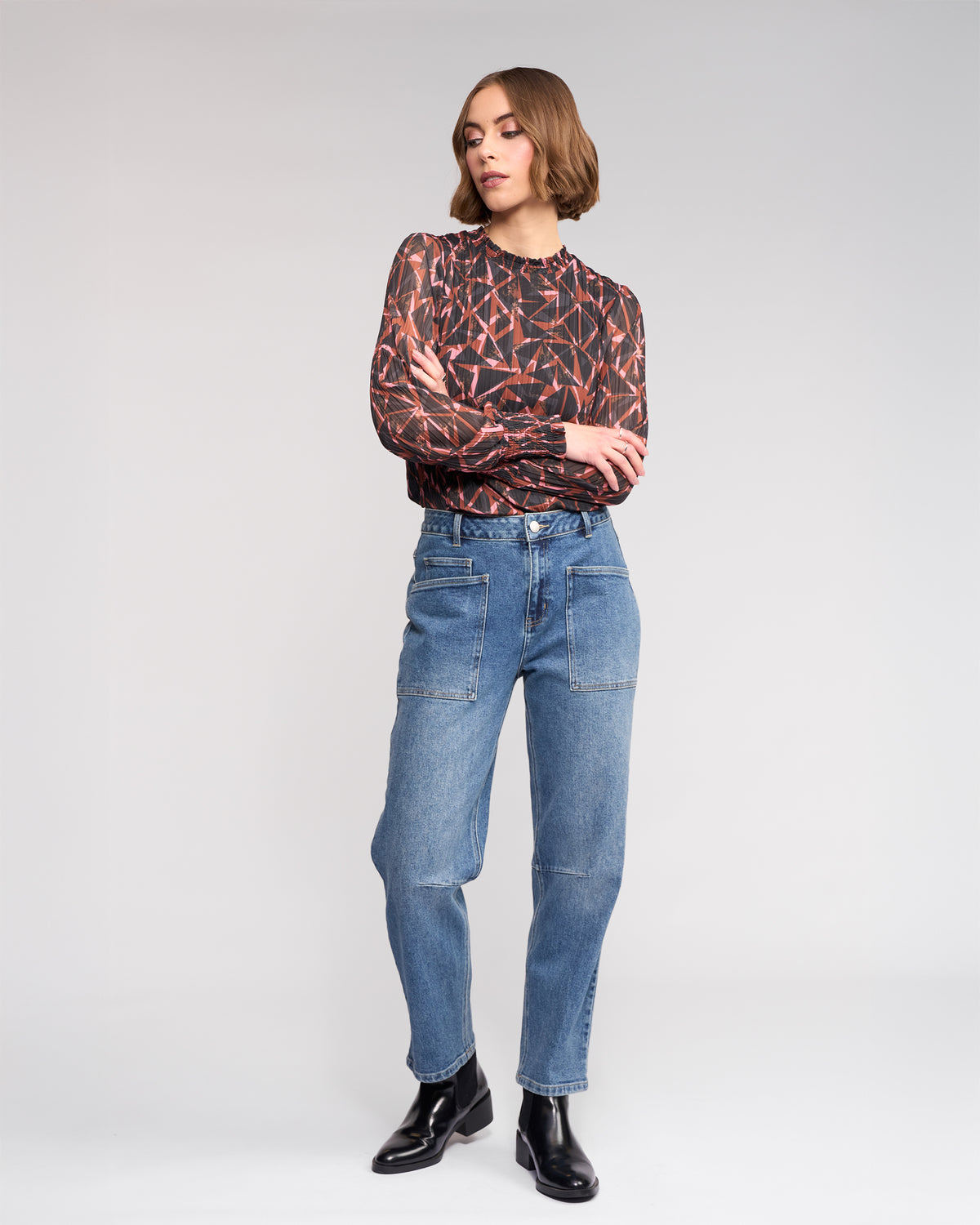A woman with short, wavy hair crosses her arms against a plain background, wearing a geometric long-sleeve top, black ankle boots, and Knewe Label’s Colt Jean in vintage denim featuring front pockets and a barrel-leg fit.