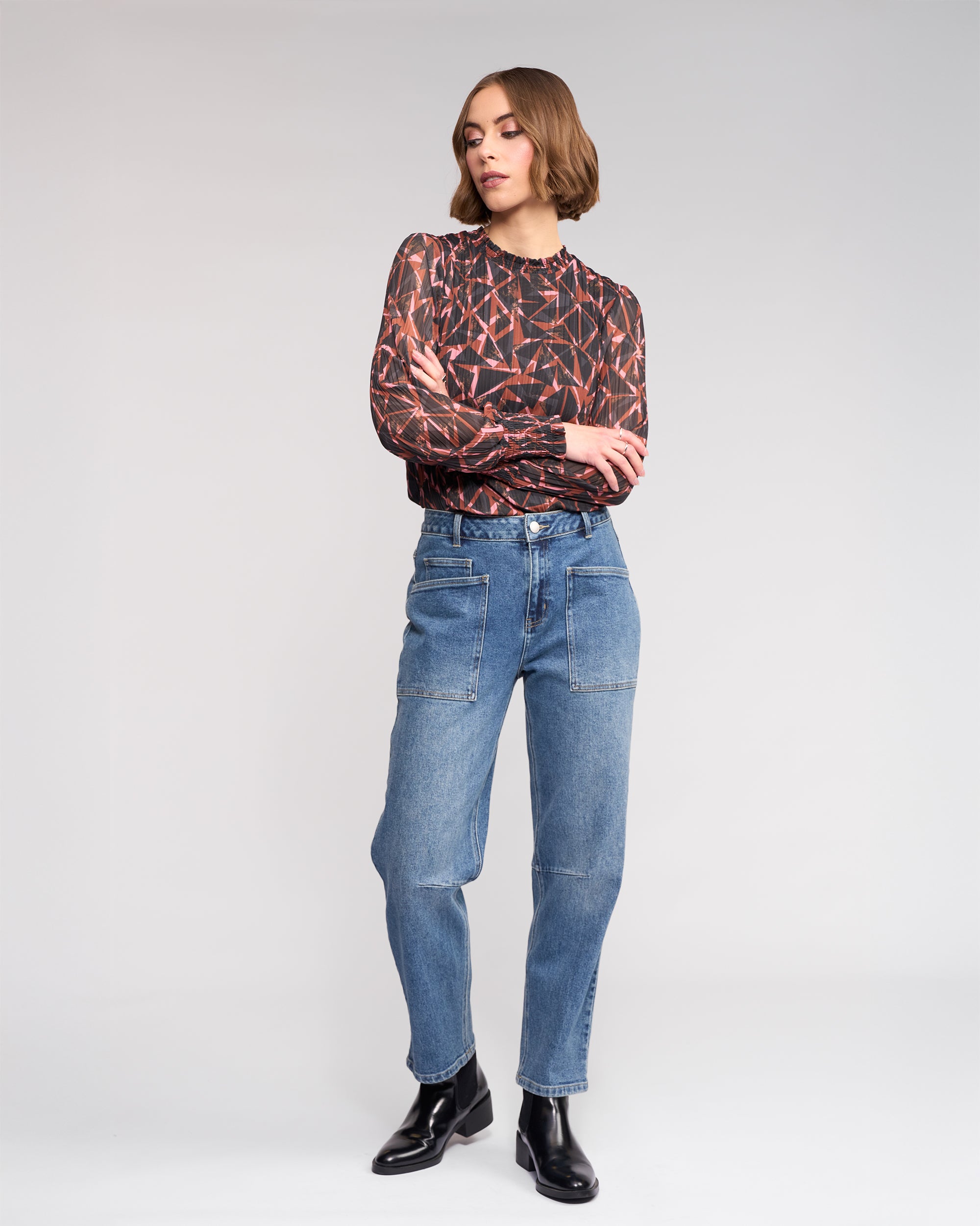 A woman with short, wavy hair crosses her arms against a plain background, wearing a geometric long-sleeve top, black ankle boots, and Knewe Label’s Colt Jean in vintage denim featuring front pockets and a barrel-leg fit.