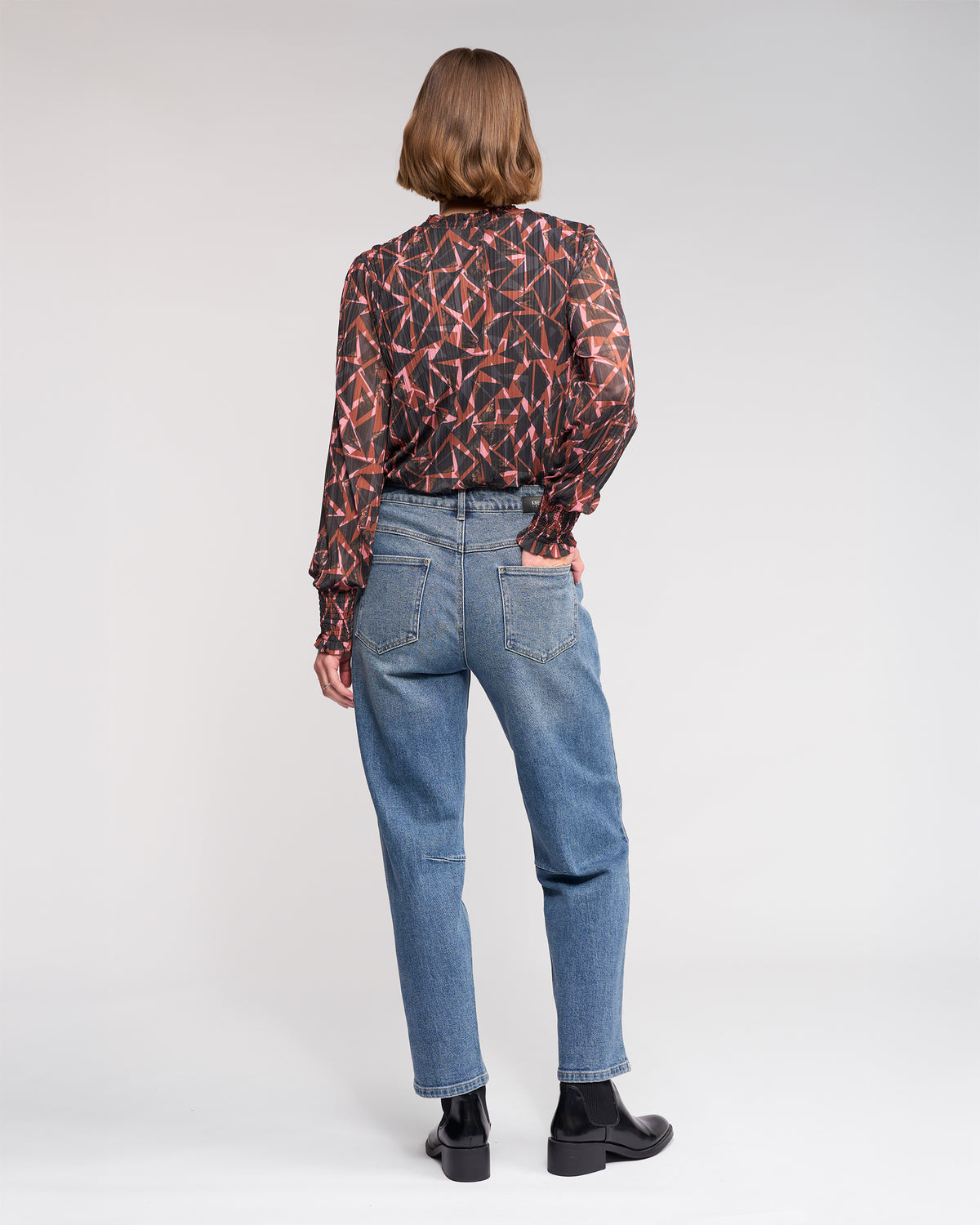 A person with short brown hair faces away, wearing a long-sleeved geometric blouse and Knewe Label's Colt Jean in Vintage Denim, paired with black shoes against a plain light background.