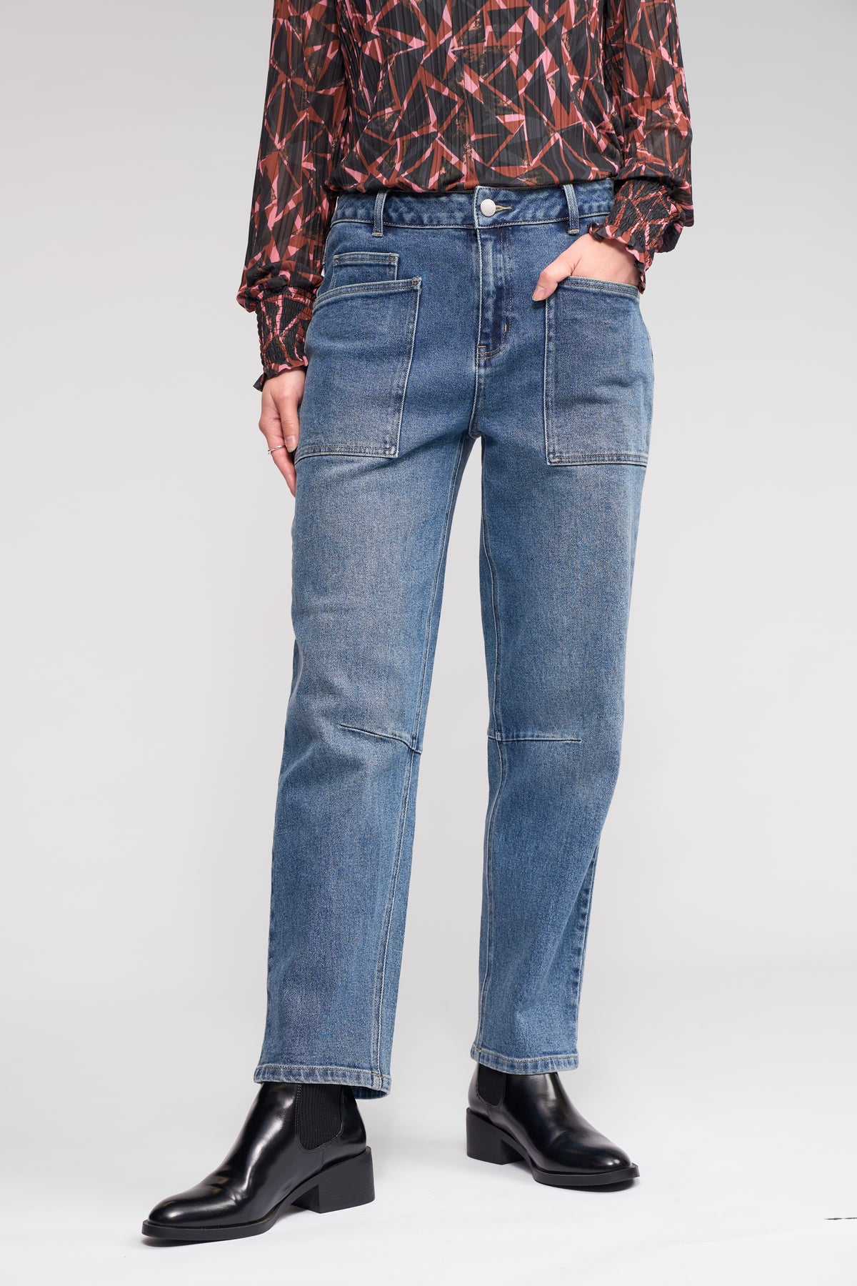 Someone wearing the Colt Jean - Vintage Denim from Knewe Label, featuring large front pockets, paired with a patterned long-sleeve blouse and black ankle boots, stands against a light background.