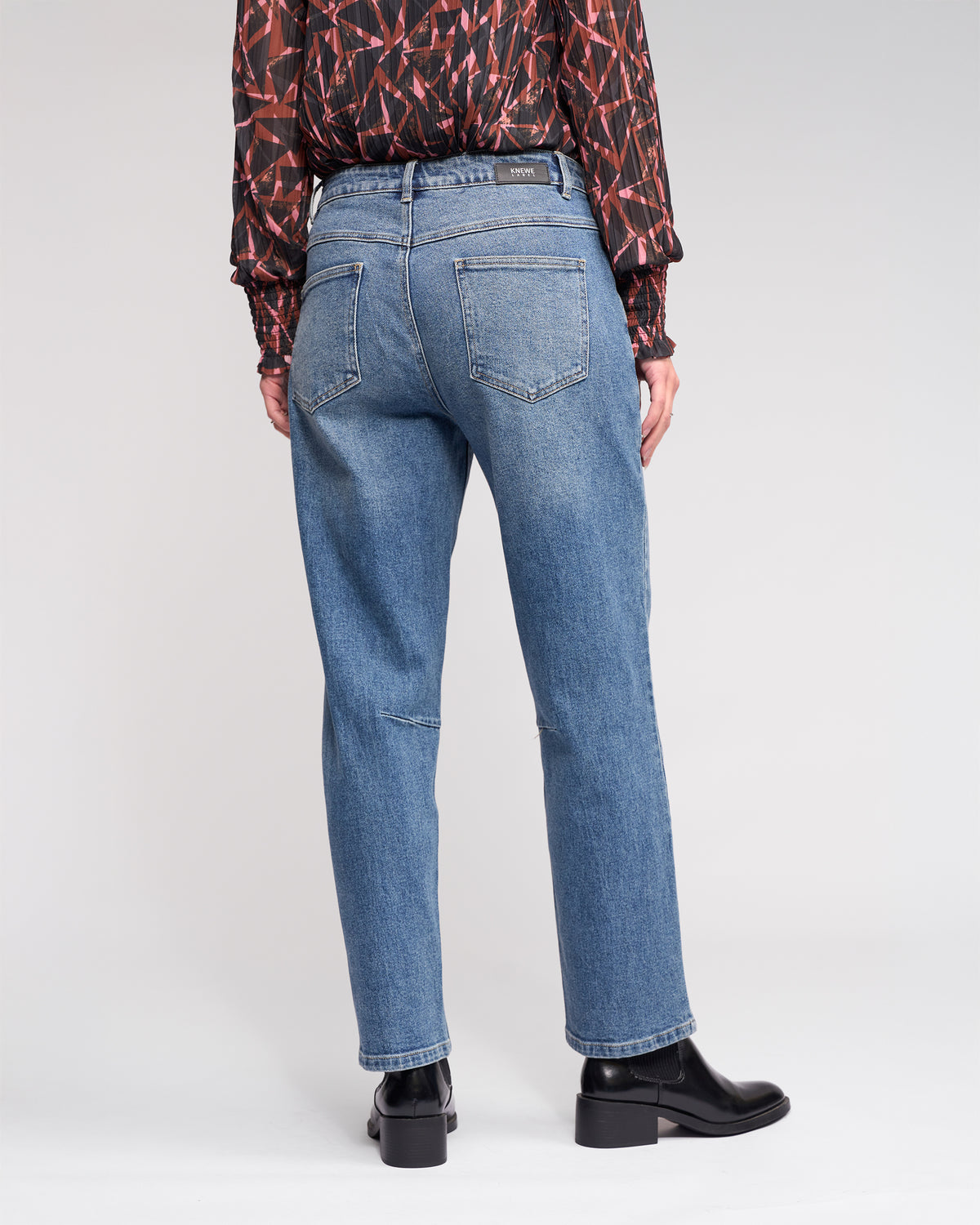 A person stands facing away, wearing Knewe Label's Colt Jean in Vintage Denim, a long-sleeved patterned red and black blouse, and black heeled ankle boots against a plain background.