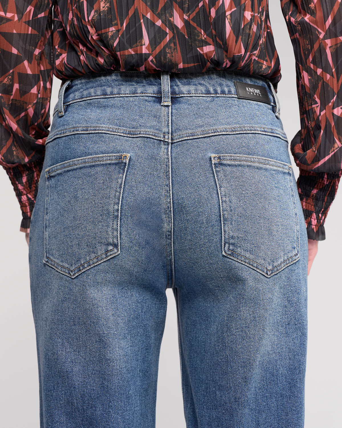 A person shown from the waist down, facing away from the camera, wears Knewe Label's Colt Jean in vintage denim and a black long-sleeve shirt with red and pink geometric patterns.