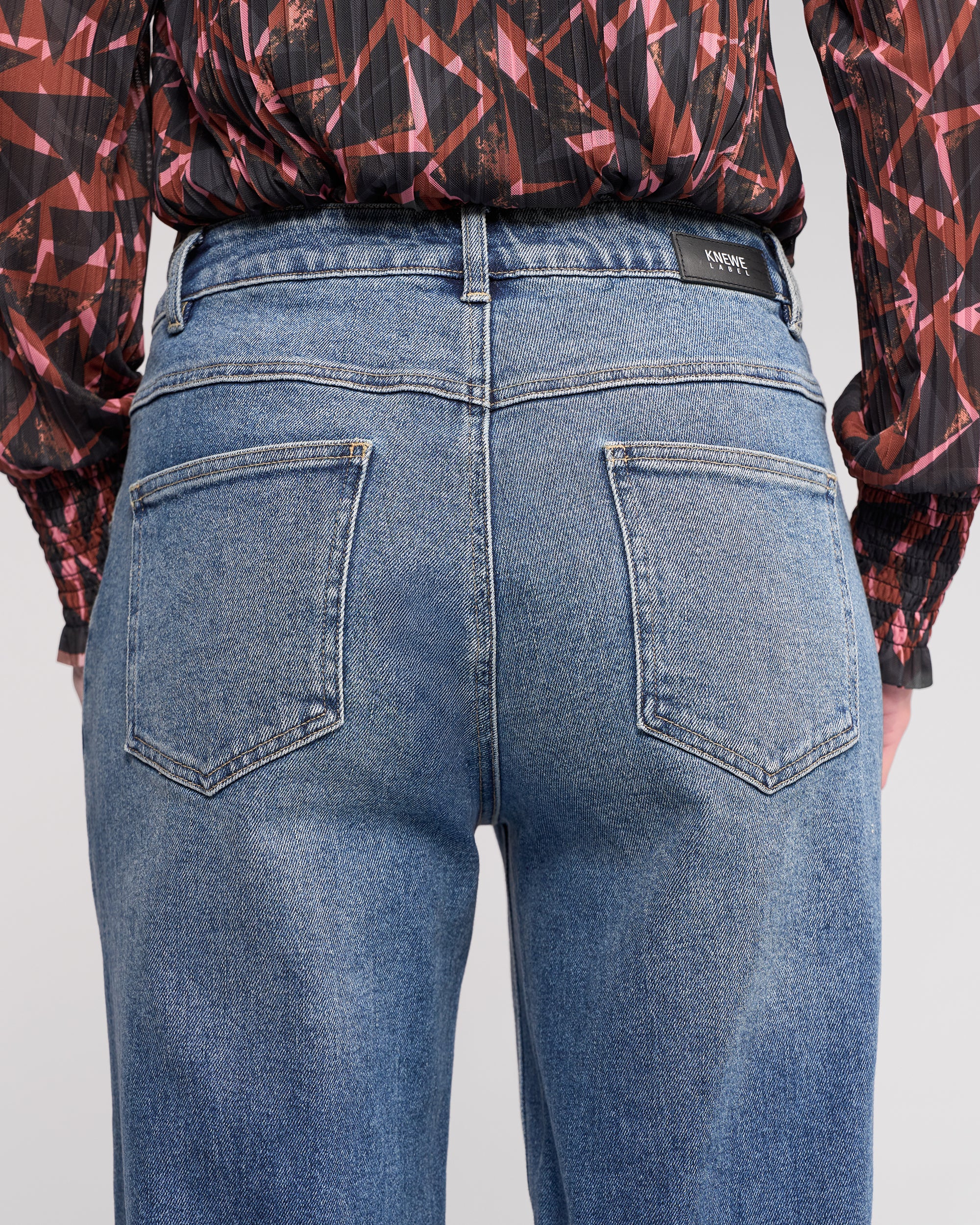 A person shown from the waist down, facing away from the camera, wears Knewe Label's Colt Jean in vintage denim and a black long-sleeve shirt with red and pink geometric patterns.