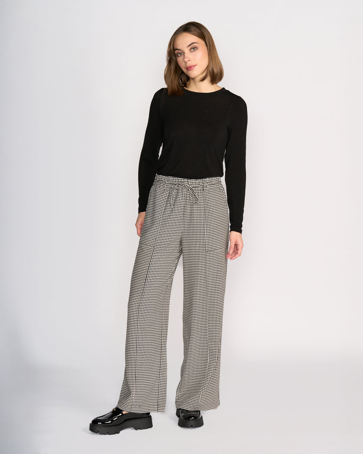 A woman with straight, shoulder-length brown hair stands against a plain background wearing Knewe Label’s Ryder Pant - Summit, a high-waisted houndstooth wide-leg style with an elasticated waistband, paired with a long-sleeve black top and black platform shoes.