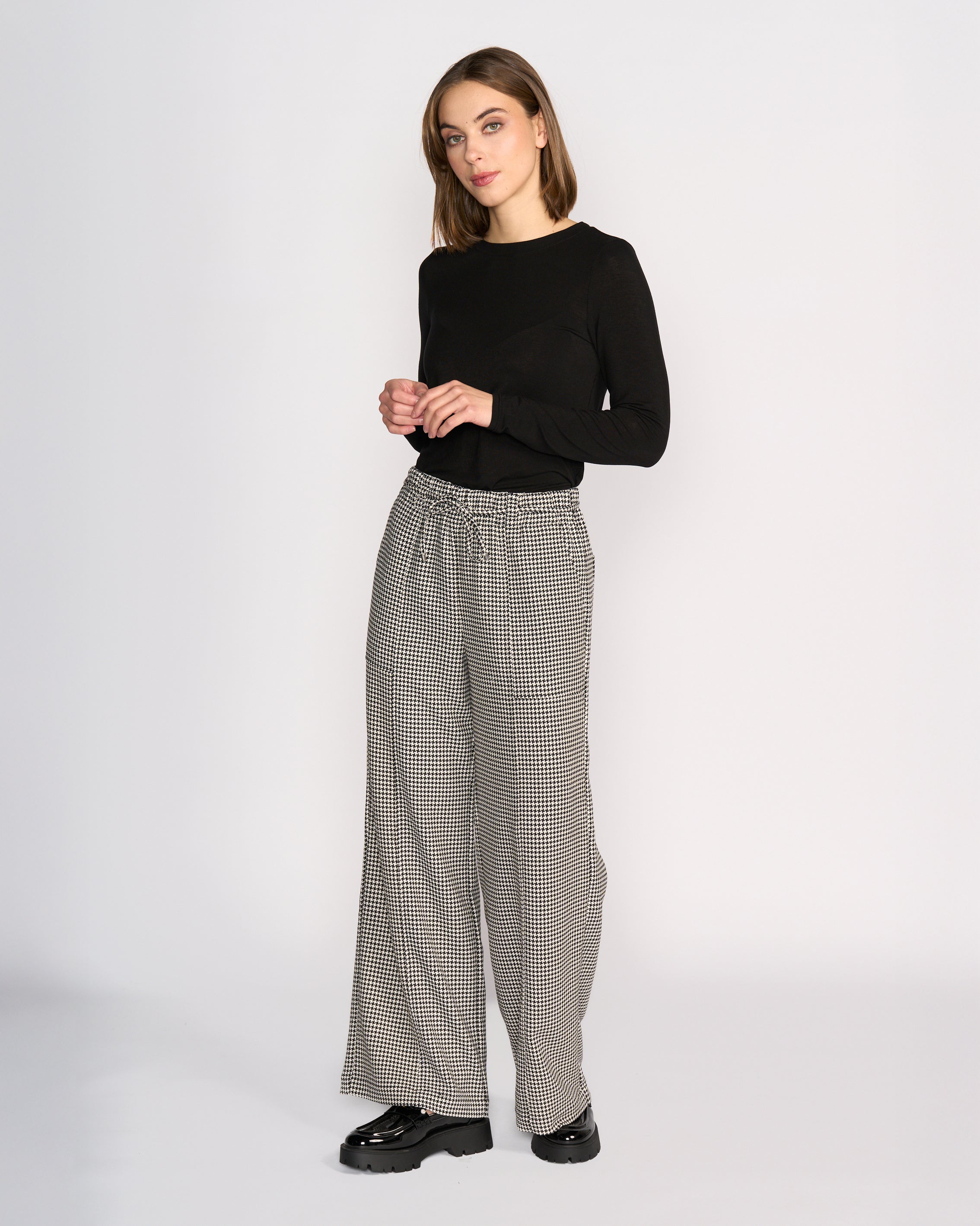 A woman stands against a plain white background, wearing a black long-sleeve top, black chunky shoes, and Knewe Label's Ryder Pant - Summit: houndstooth wide-leg pants with an elasticated waistband.