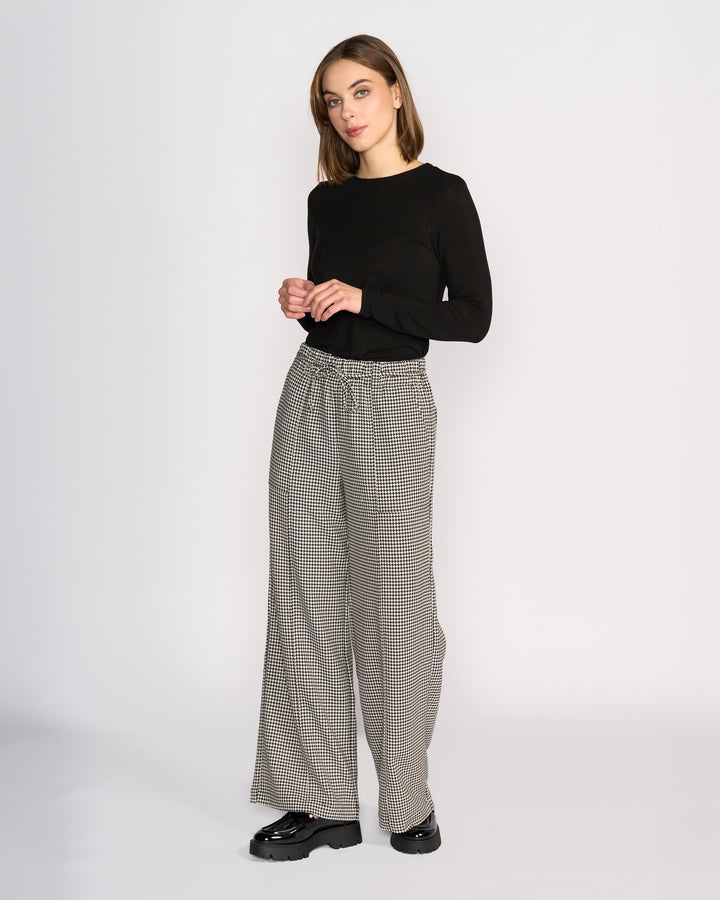 A woman stands against a plain white background, wearing a black long-sleeve top, black chunky shoes, and Knewe Label's Ryder Pant - Summit: houndstooth wide-leg pants with an elasticated waistband.