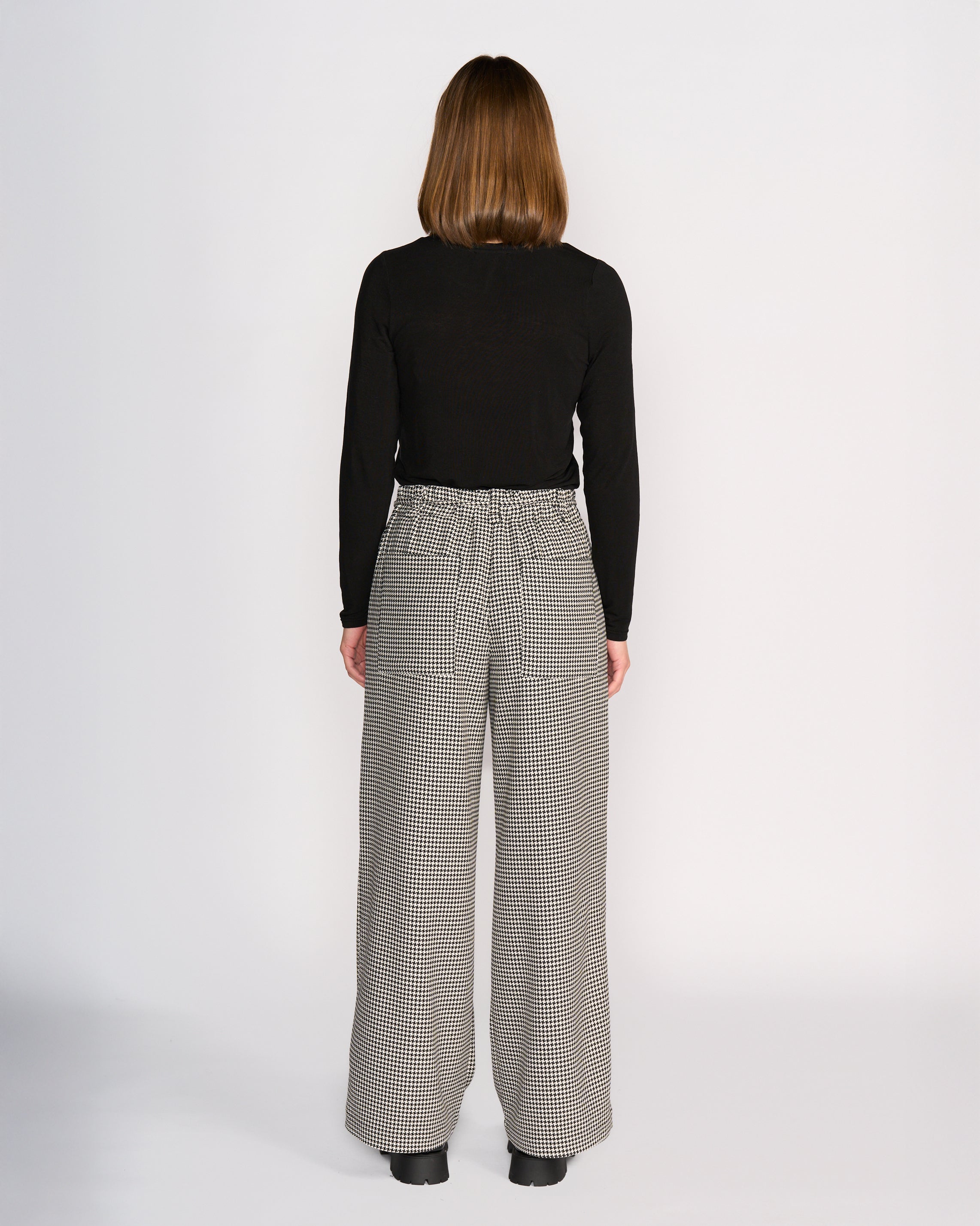 A person with straight, shoulder-length brown hair stands facing away, wearing a black long-sleeve top and Knewe Label Ryder Pant - Summit, featuring a high-waisted, wide-leg houndstooth design with an elastic waistband and black shoes against a plain white background.
