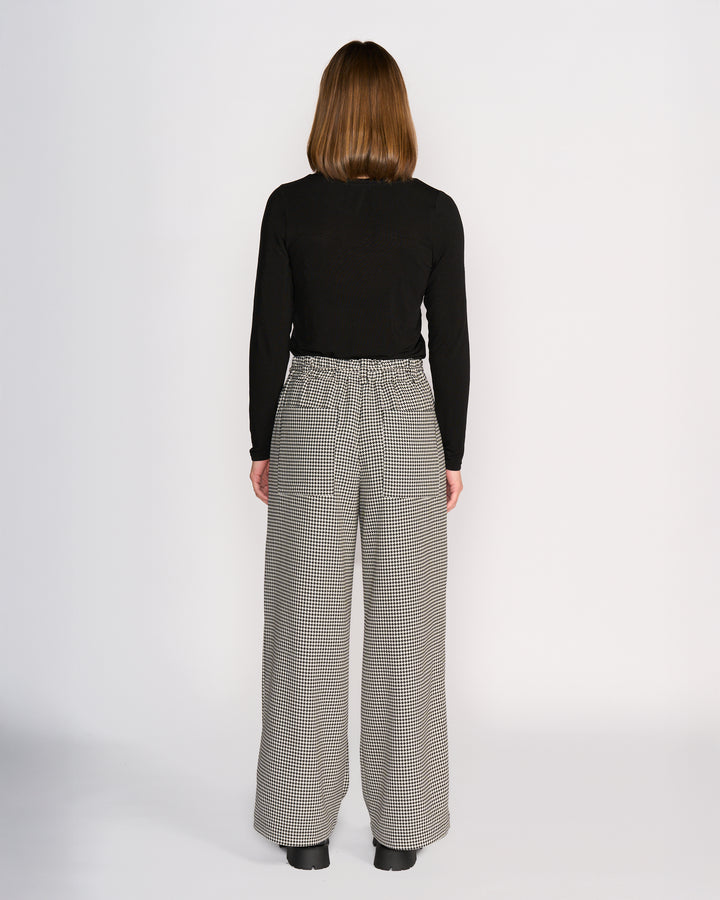 A person with straight, shoulder-length brown hair stands facing away, wearing a black long-sleeve top and Knewe Label Ryder Pant - Summit, featuring a high-waisted, wide-leg houndstooth design with an elastic waistband and black shoes against a plain white background.