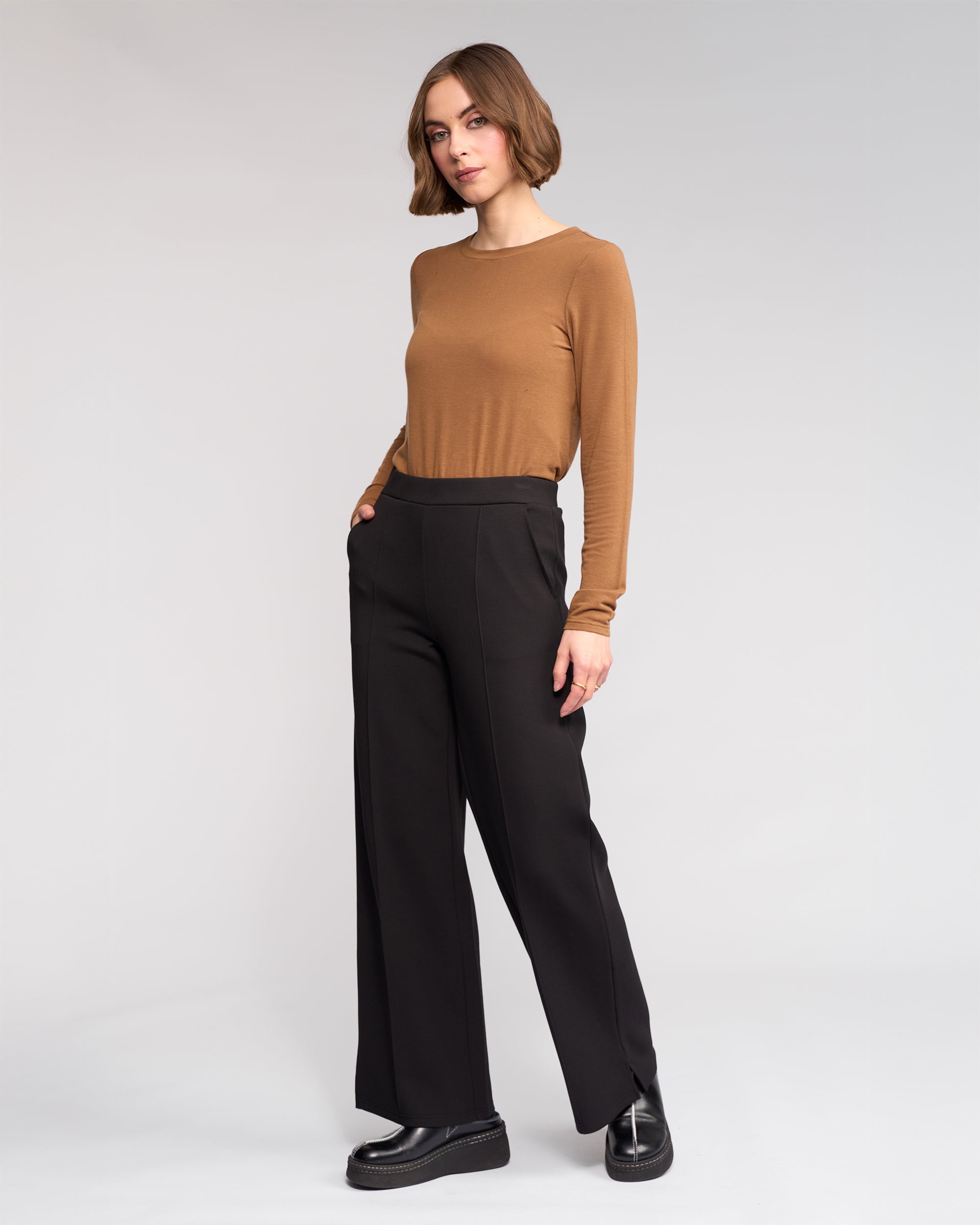 A woman with short brown hair wears the Knewe Label Dune Pant in Black—stretchy, wide-leg pants—paired with a fitted brown long-sleeve top and black platform shoes, standing with one hand in her pocket against a plain light background.