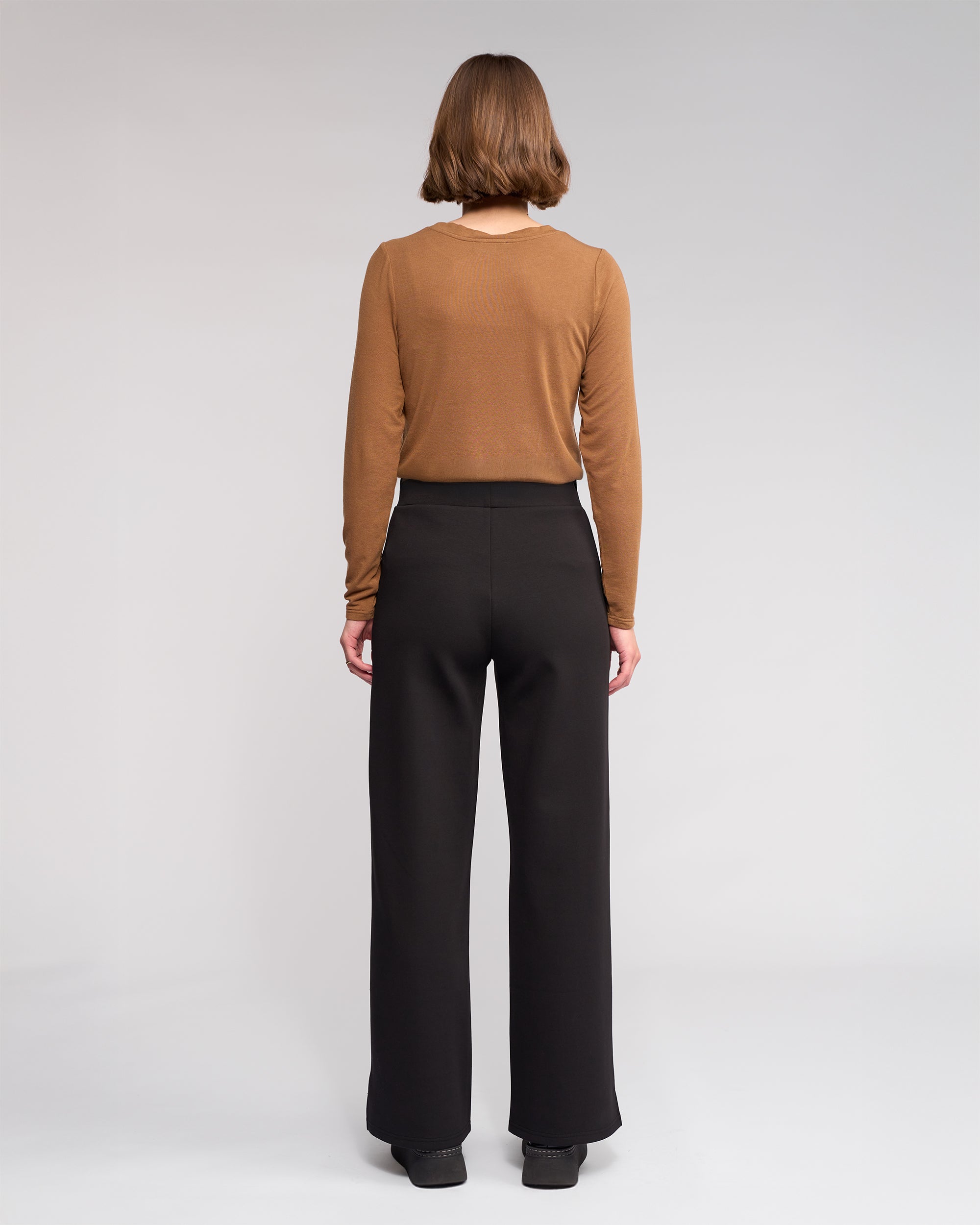 A person with short brown hair faces away from the camera, wearing a long-sleeve brown top, Knewe Label’s Dune Pant in black, and black shoes in a studio with a neutral background.