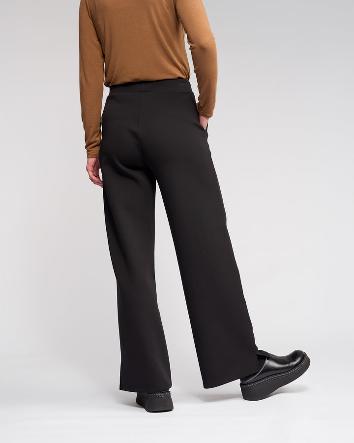 A person wearing the Knewe Label Dune Pant in black, paired with a brown long-sleeve top and black platform shoes, stands with their back to the camera against a plain light background.