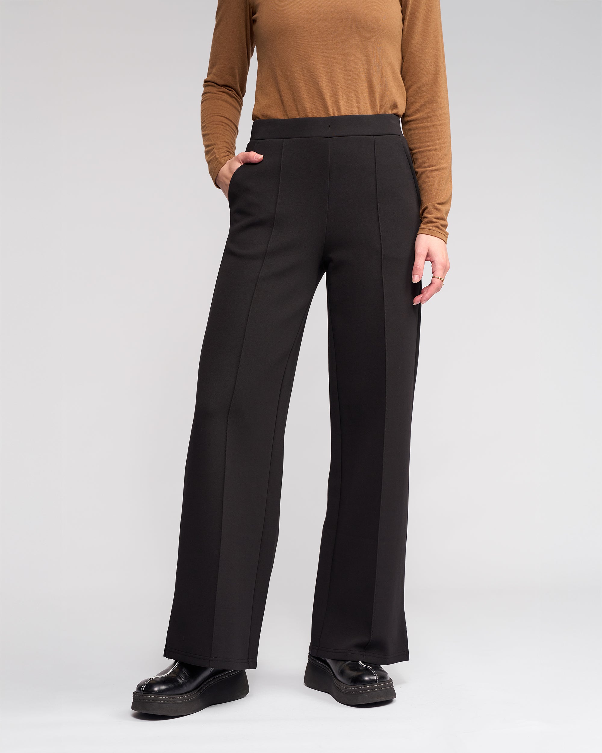 A person stands against a light gray background wearing Knewe Label’s Dune Pant in black, paired with a long-sleeve brown top and black platform shoes. Their left hand is tucked into the pocket of the high-waisted, wide-leg pants.