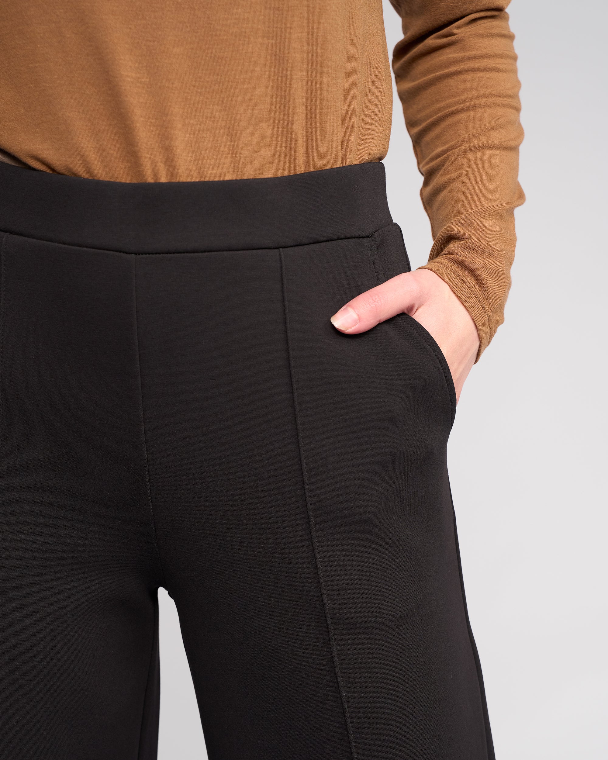 A model wearing Knewe Label's Dune Pant in Black with a brown long-sleeve top, one hand in the pocket, shown from shoulders to mid-thigh against a plain light background.