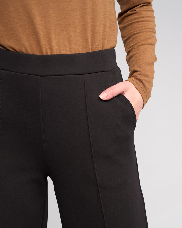A model wearing Knewe Label's Dune Pant in Black with a brown long-sleeve top, one hand in the pocket, shown from shoulders to mid-thigh against a plain light background.