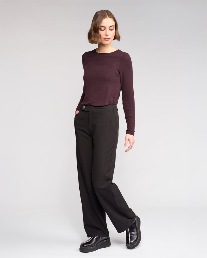 A person with a short bob wears the Knewe Label Blaze Pant in black, styled with a long-sleeve maroon top and black platform shoes, standing against a plain light background, hand in pocket and looking down.