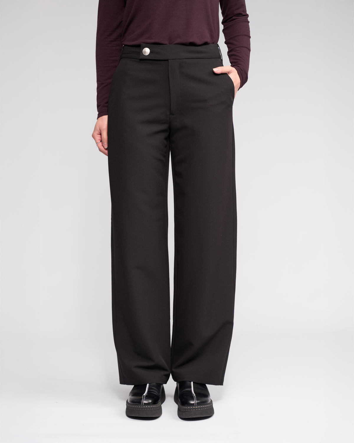 Someone wearing the Knewe Label Blaze Pant in Black stands with one hand in their pocket, paired with a dark long-sleeve top and black shoes, against a plain light background.