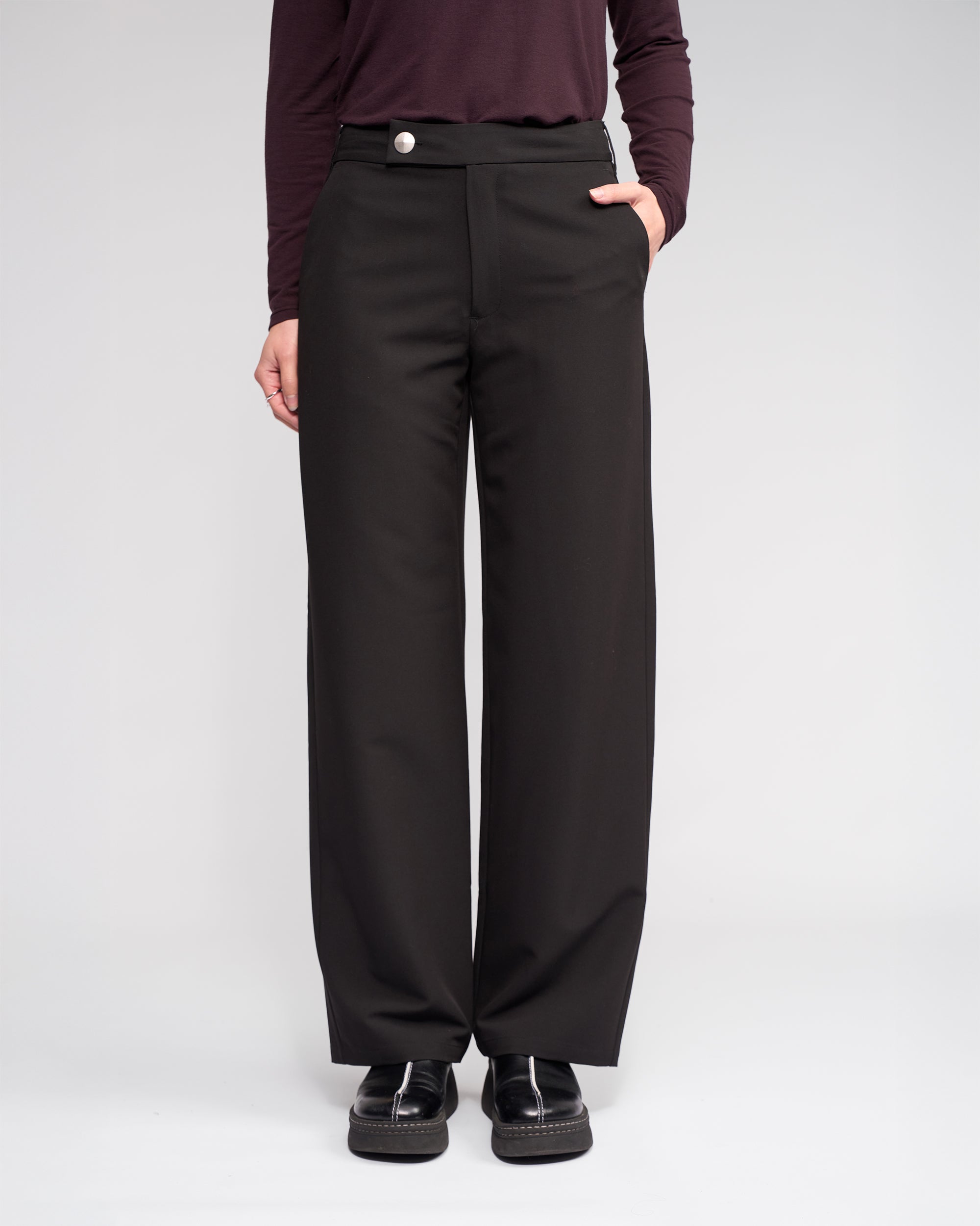 Someone wearing the Knewe Label Blaze Pant in Black stands with one hand in their pocket, paired with a dark long-sleeve top and black shoes, against a plain light background.