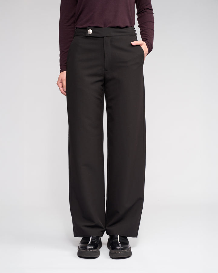 Someone wearing the Knewe Label Blaze Pant in Black stands with one hand in their pocket, paired with a dark long-sleeve top and black shoes, against a plain light background.