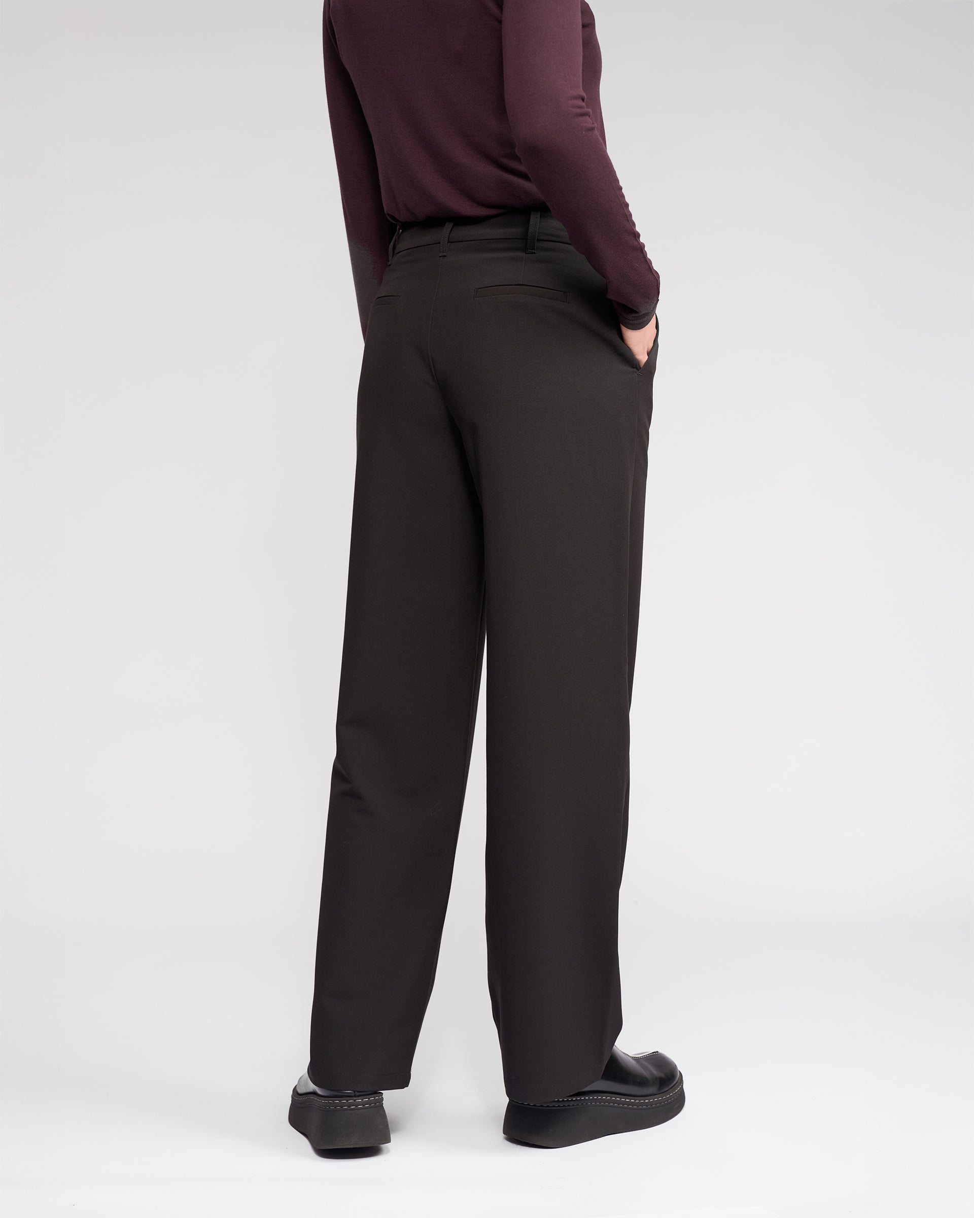 Facing away, a person wears the Knewe Label Blaze Pant in black, featuring a wide-leg fit and asymmetric waistband. They pair it with a long-sleeve burgundy top, hand in pocket, set against a plain white background for a formal look.