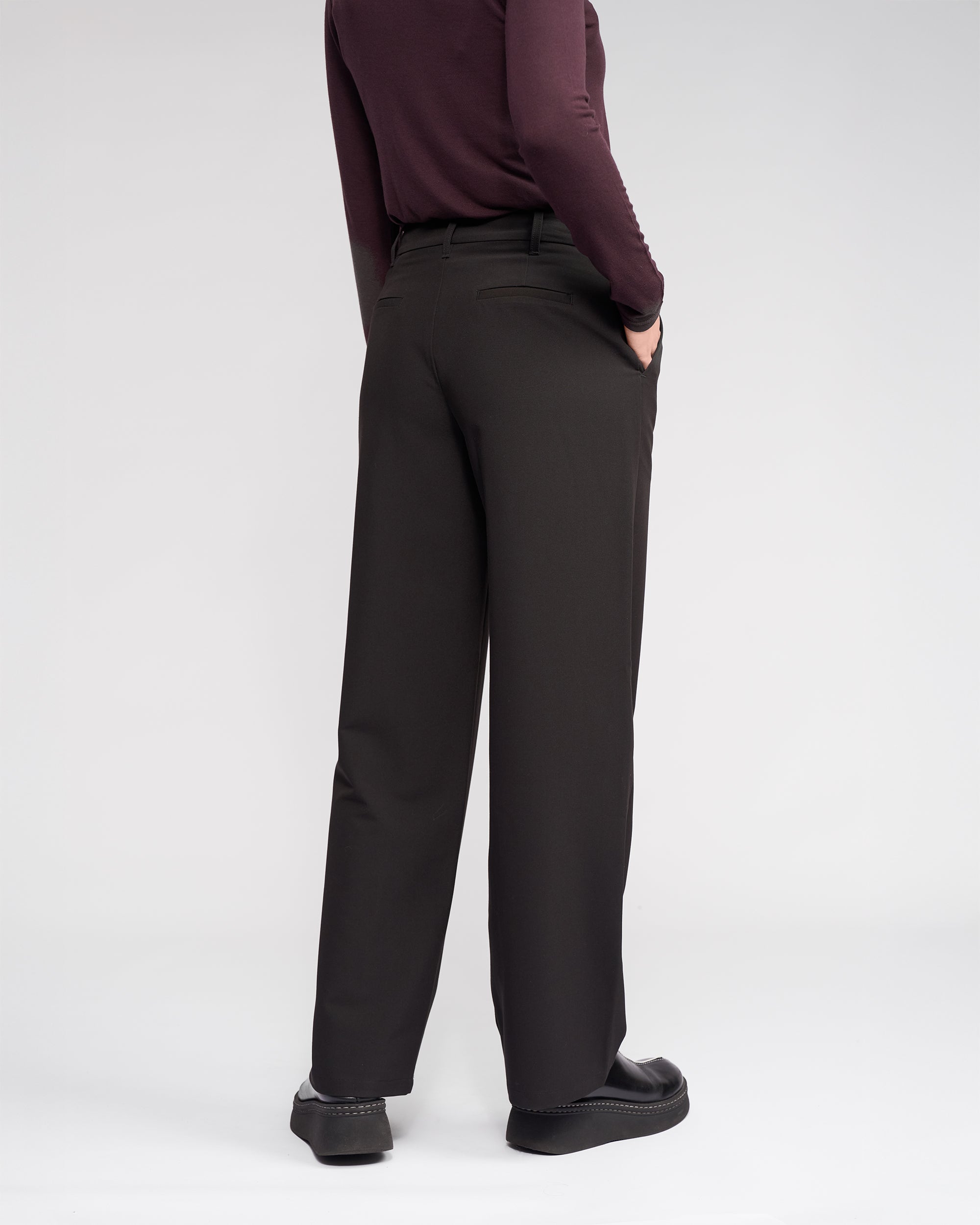Facing away, a person wears the Knewe Label Blaze Pant in black, featuring a wide-leg fit and asymmetric waistband. They pair it with a long-sleeve burgundy top, hand in pocket, set against a plain white background for a formal look.