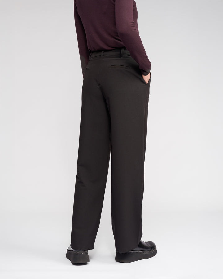 Facing away, a person wears the Knewe Label Blaze Pant in black, featuring a wide-leg fit and asymmetric waistband. They pair it with a long-sleeve burgundy top, hand in pocket, set against a plain white background for a formal look.