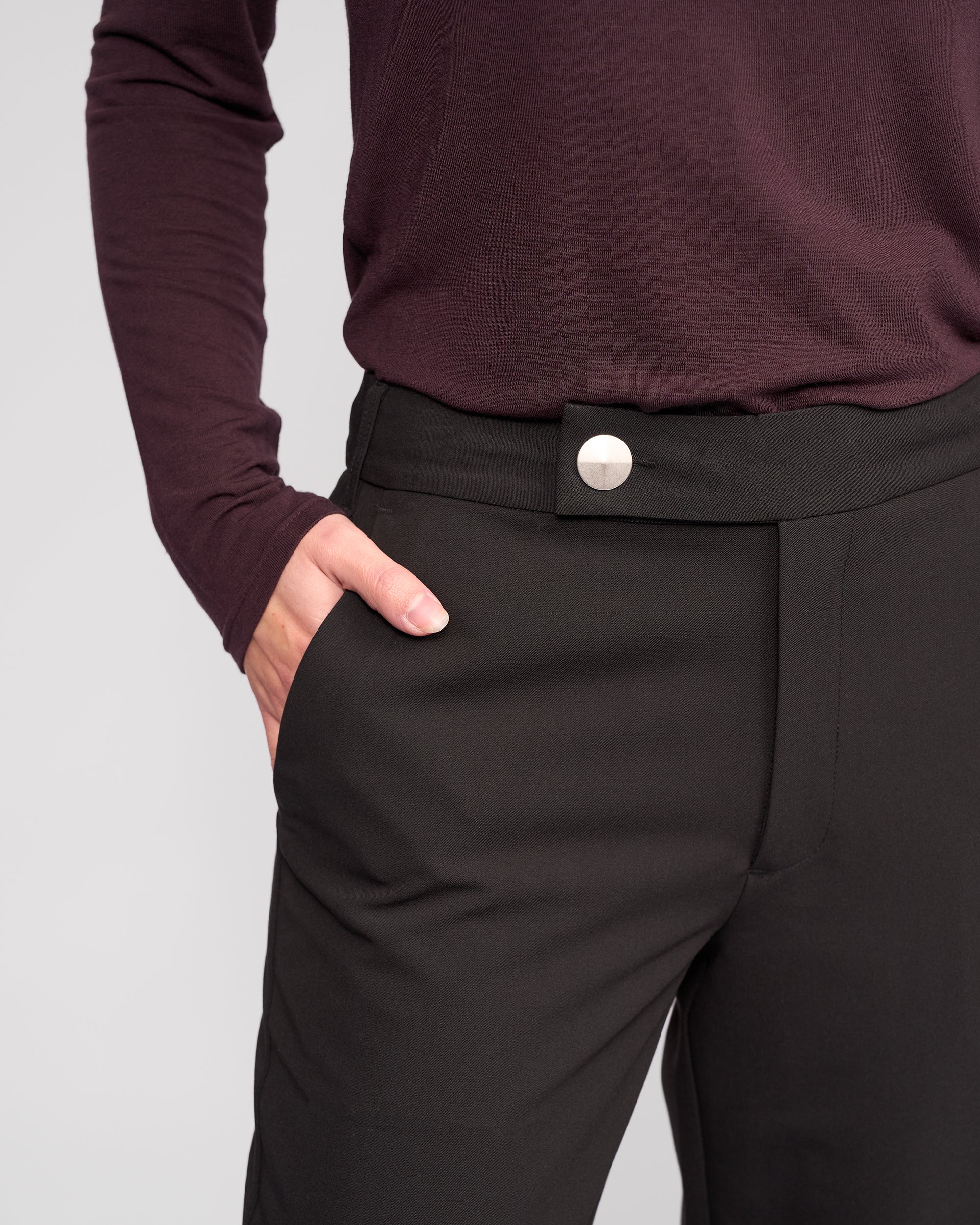 A person wearing a long-sleeve maroon top and Knewe Label's Blaze Pant in black, featuring a silver button, stands with one hand in their pocket against a plain light background.