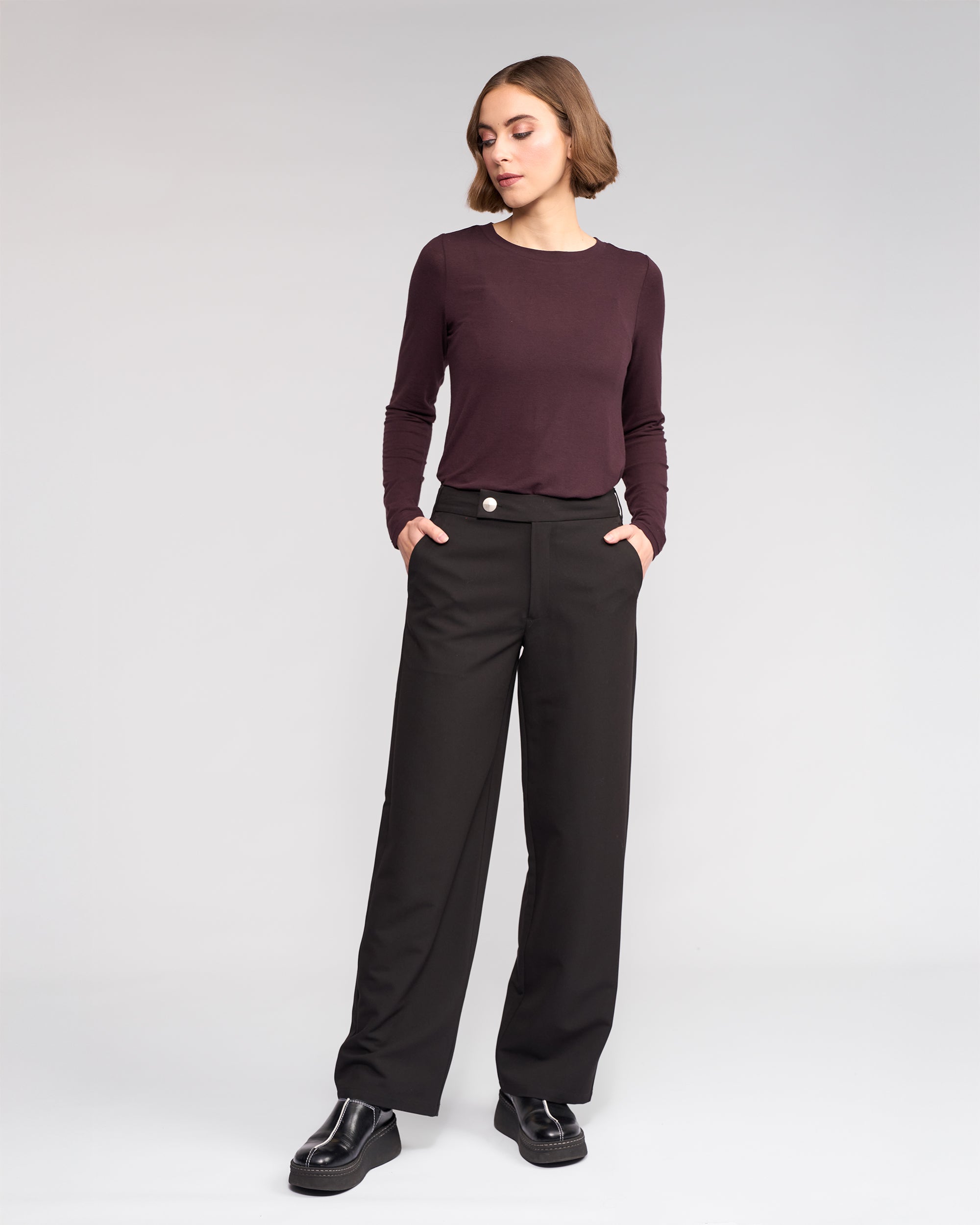 A woman wears the Knewe Label Blaze Pant in black, featuring a wide leg and asymmetric waistband, paired with a long-sleeve burgundy top; she stands with hands in her pockets against a plain light gray background.