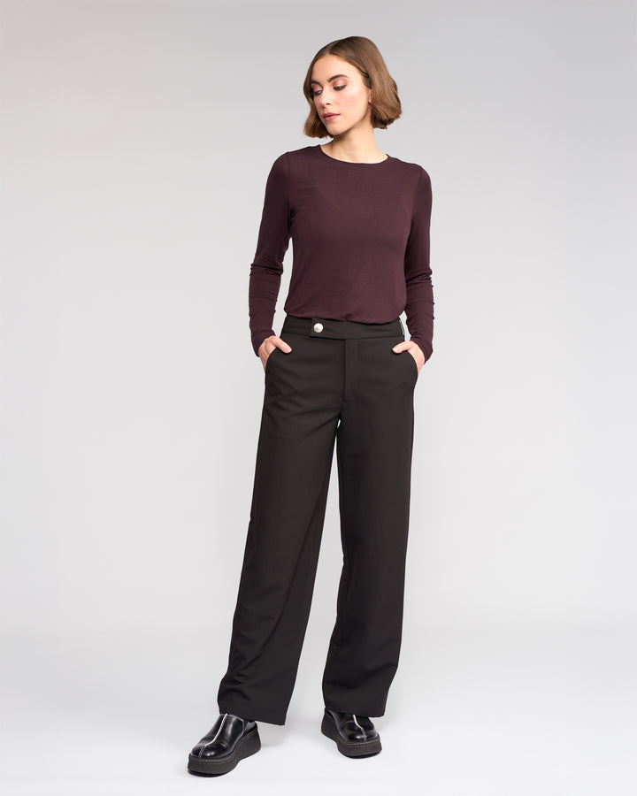 A woman wears the Knewe Label Blaze Pant in black, featuring a wide leg and asymmetric waistband, paired with a long-sleeve burgundy top; she stands with hands in her pockets against a plain light gray background.