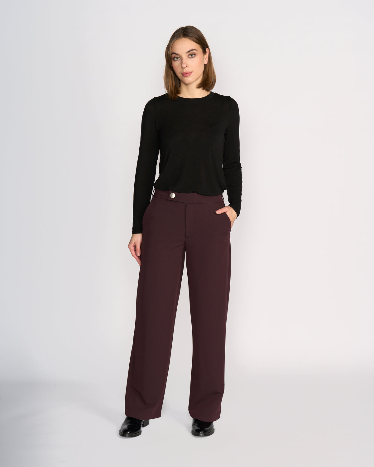A woman stands against a plain white background wearing the Knewe Label Blaze Pant in Raisin and a black long-sleeve top. With one hand in her pocket, she exudes effortless style and confidence in this polished, formal look.