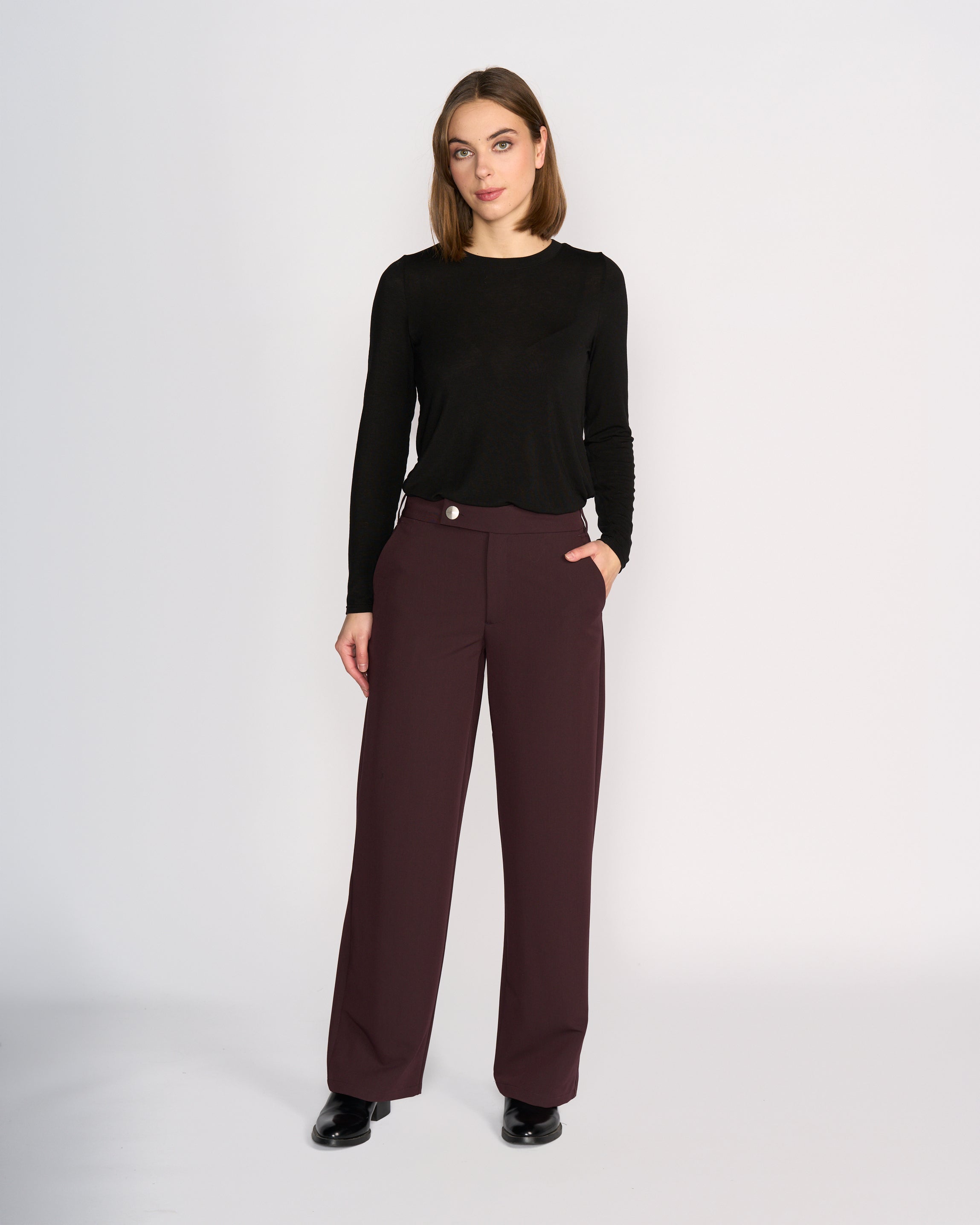 A woman stands against a plain white background wearing the Knewe Label Blaze Pant in Raisin and a black long-sleeve top. With one hand in her pocket, she exudes effortless style and confidence in this polished, formal look.