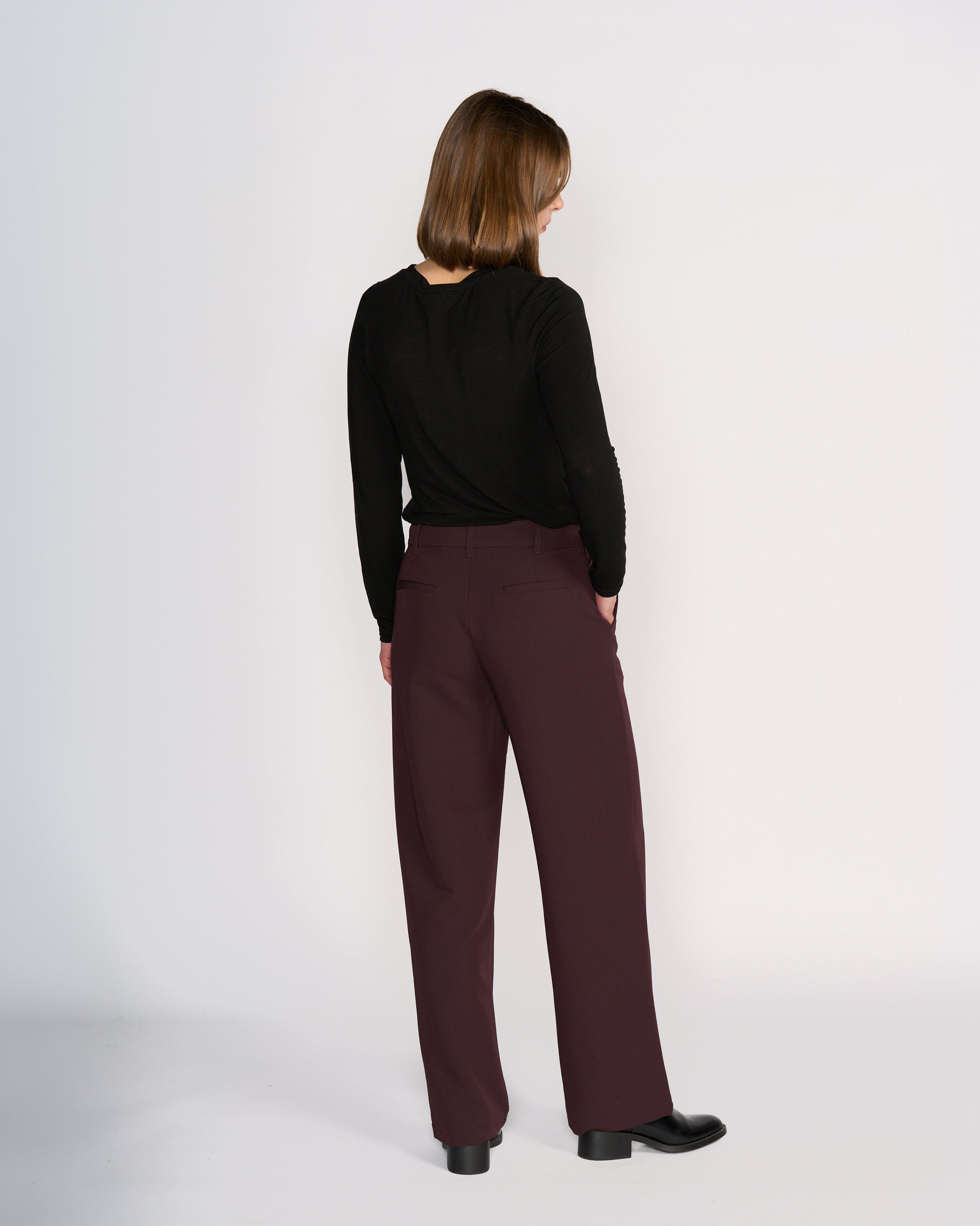 A woman faces away from the camera wearing Knewe Label's Blaze Pant in Raisin, styled with a long-sleeved black top and black shoes, creating a formal silhouette against a plain white background.