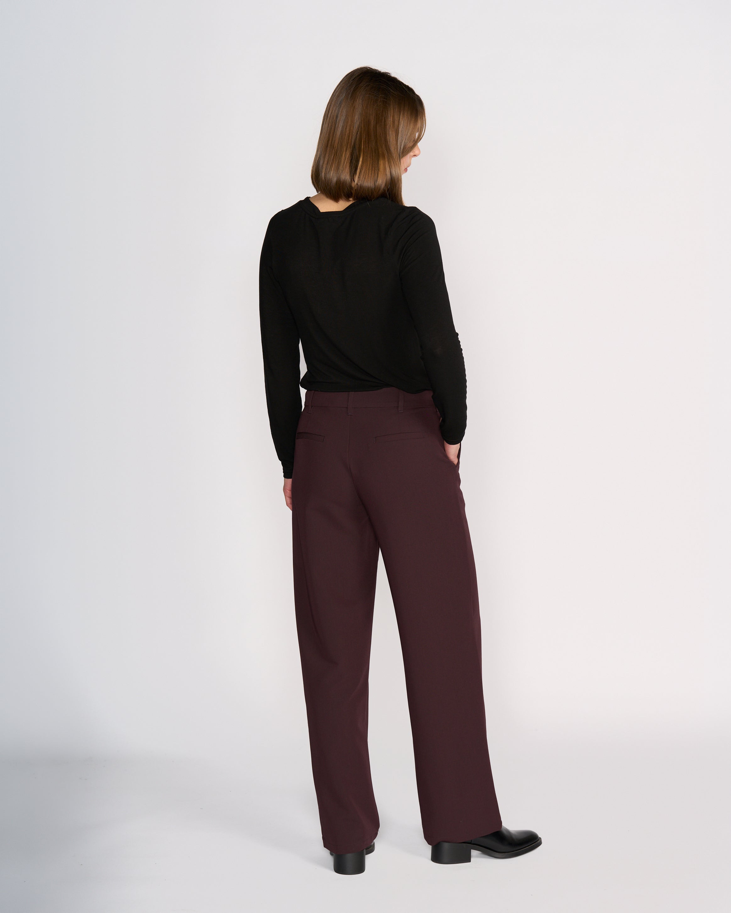A woman faces away from the camera wearing Knewe Label's Blaze Pant in Raisin, styled with a long-sleeved black top and black shoes, creating a formal silhouette against a plain white background.