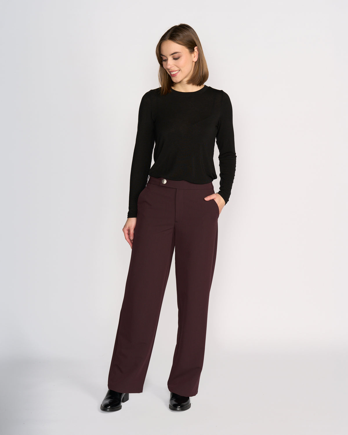 A woman models Knewe Label's Blaze Pant in Raisin, paired with a black long-sleeve top and black shoes. She stands against a plain light background, hand in pocket, smiling and looking down to the side with effortless tailored style.