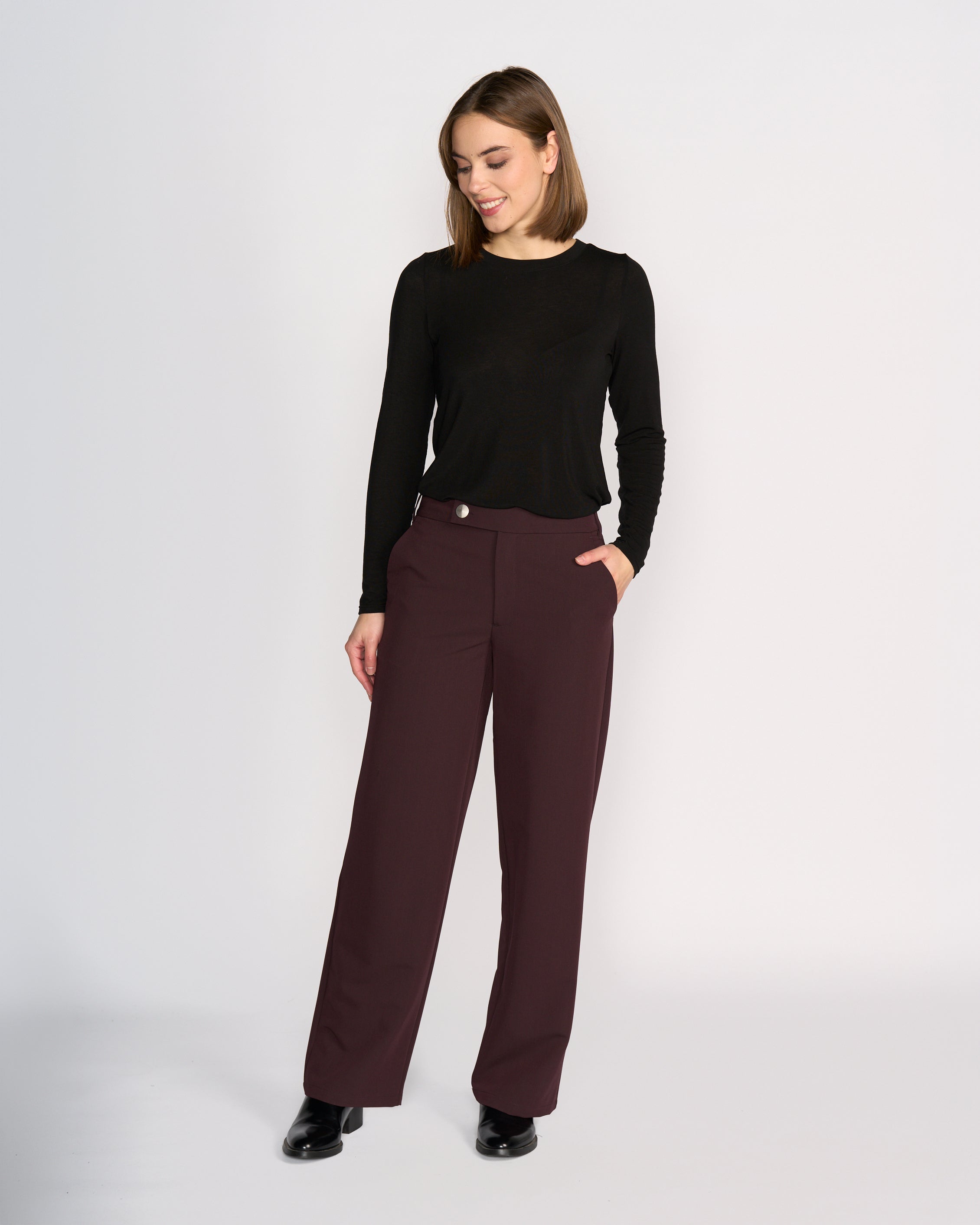 A woman models Knewe Label's Blaze Pant in Raisin, paired with a black long-sleeve top and black shoes. She stands against a plain light background, hand in pocket, smiling and looking down to the side with effortless tailored style.