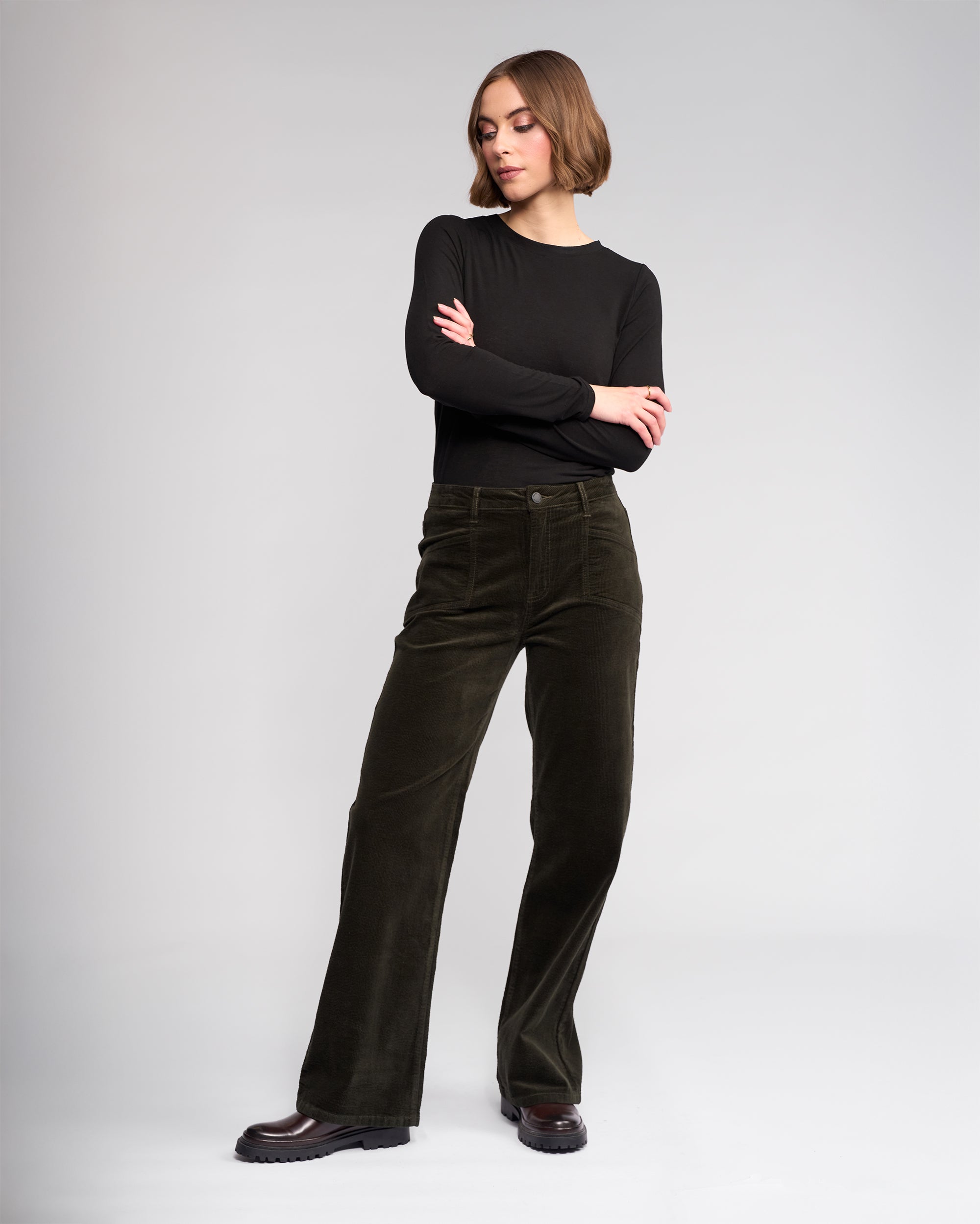 A woman stands against a light gray background, wearing a long-sleeve black top, black shoes, and Knewe Label's Dallas Cord - Hunter wide-leg pants in dark green, with her arms crossed and looking to the side.