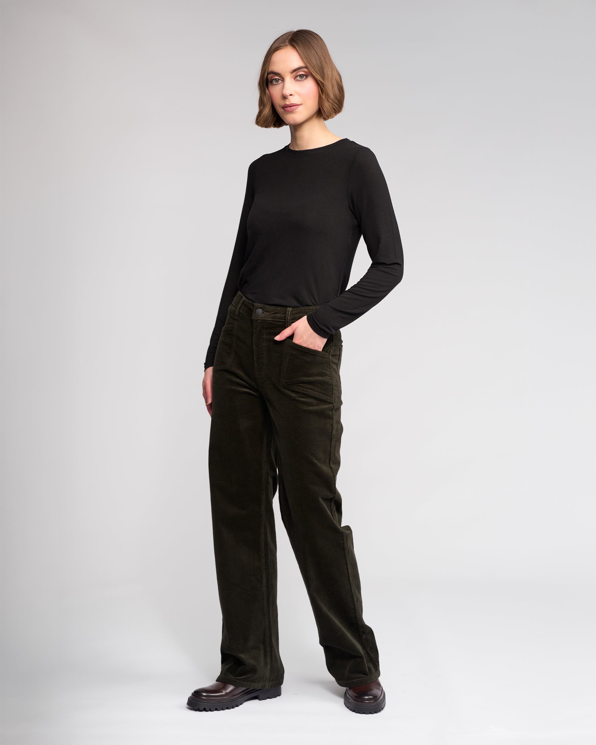 A woman stands against a plain background wearing the Knewe Label Dallas Cord – Hunter dark green wide-leg pants, a black long-sleeve shirt, and dark shoes. She has a neutral expression and one hand in her pocket.
