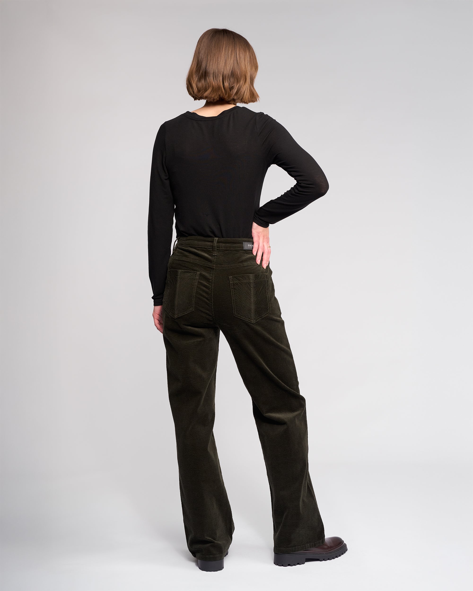 A person with short light brown hair stands facing away, wearing the Knewe Label Dallas Cord - Hunter dark green full-length pants and a black long-sleeve shirt against a plain light gray background. One hand rests on their hip.
